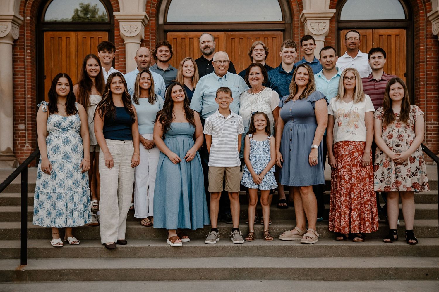 Large family gathering posed on brick church steps wearing coordinated blue and floral outfits.