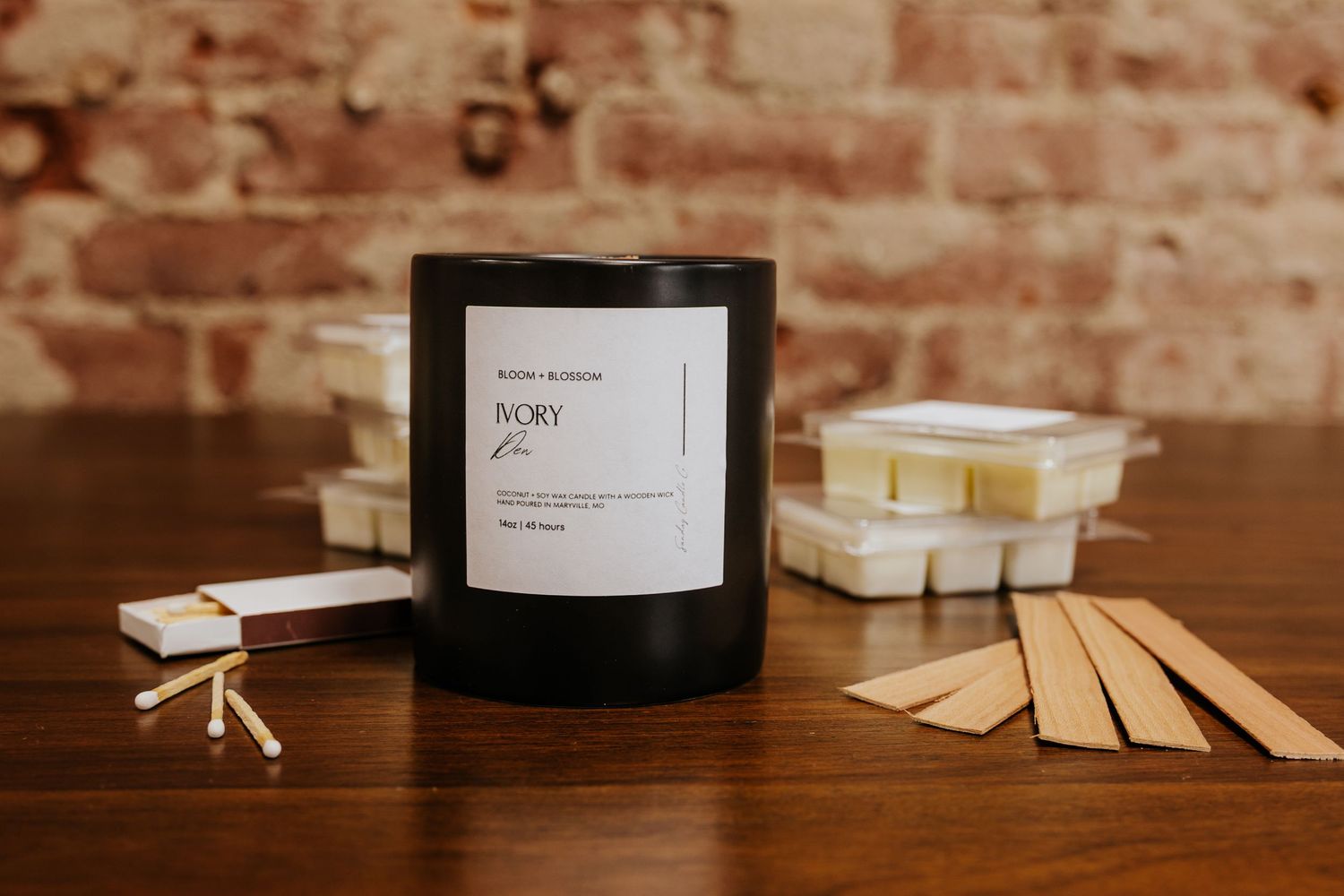 Black IVORY candle jar beside wax melts and wooden wicks on a wooden table with a brick wall background.