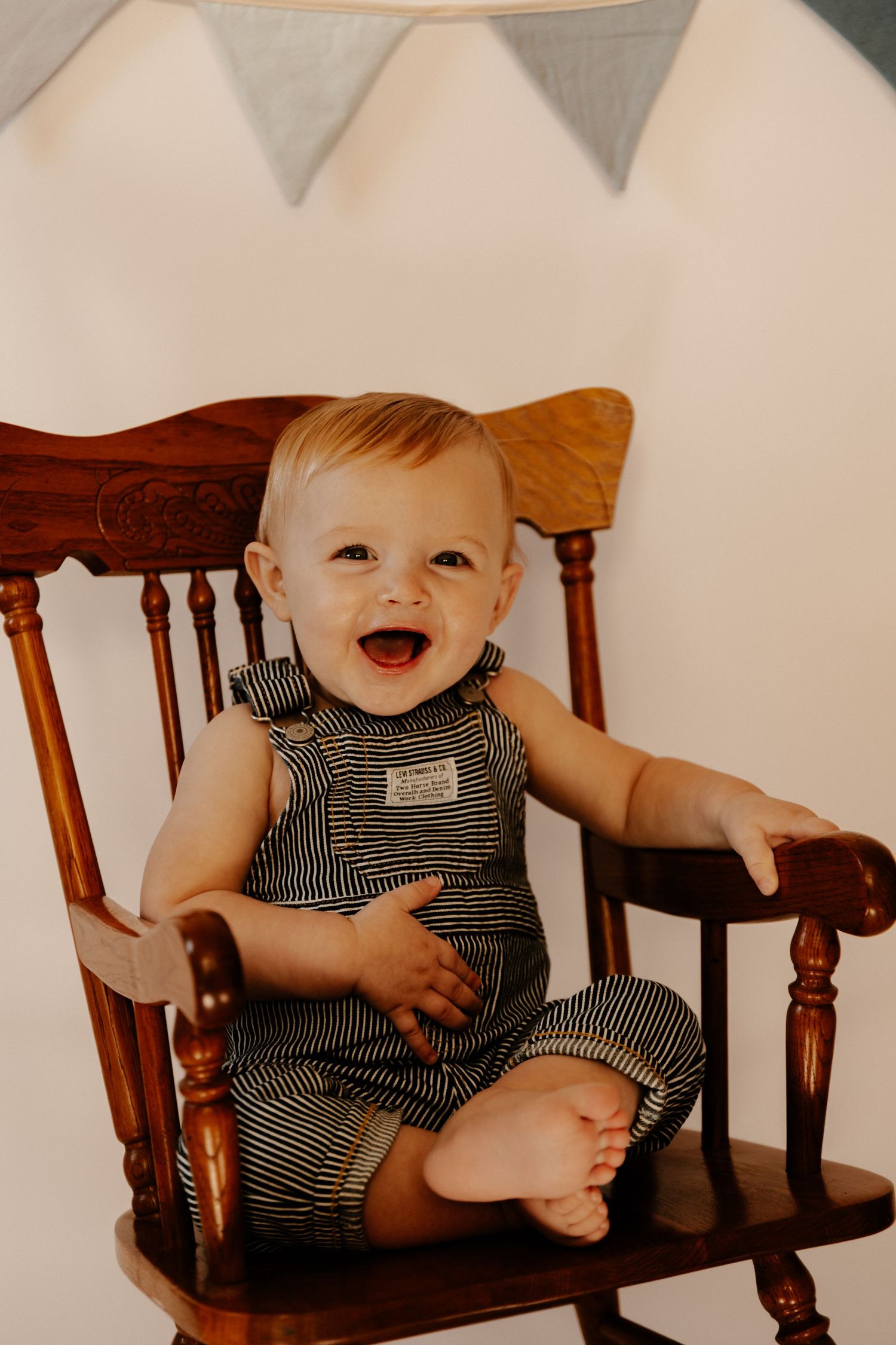 Smiling baby wearing striped overalls sitting in wooden rocking chair.