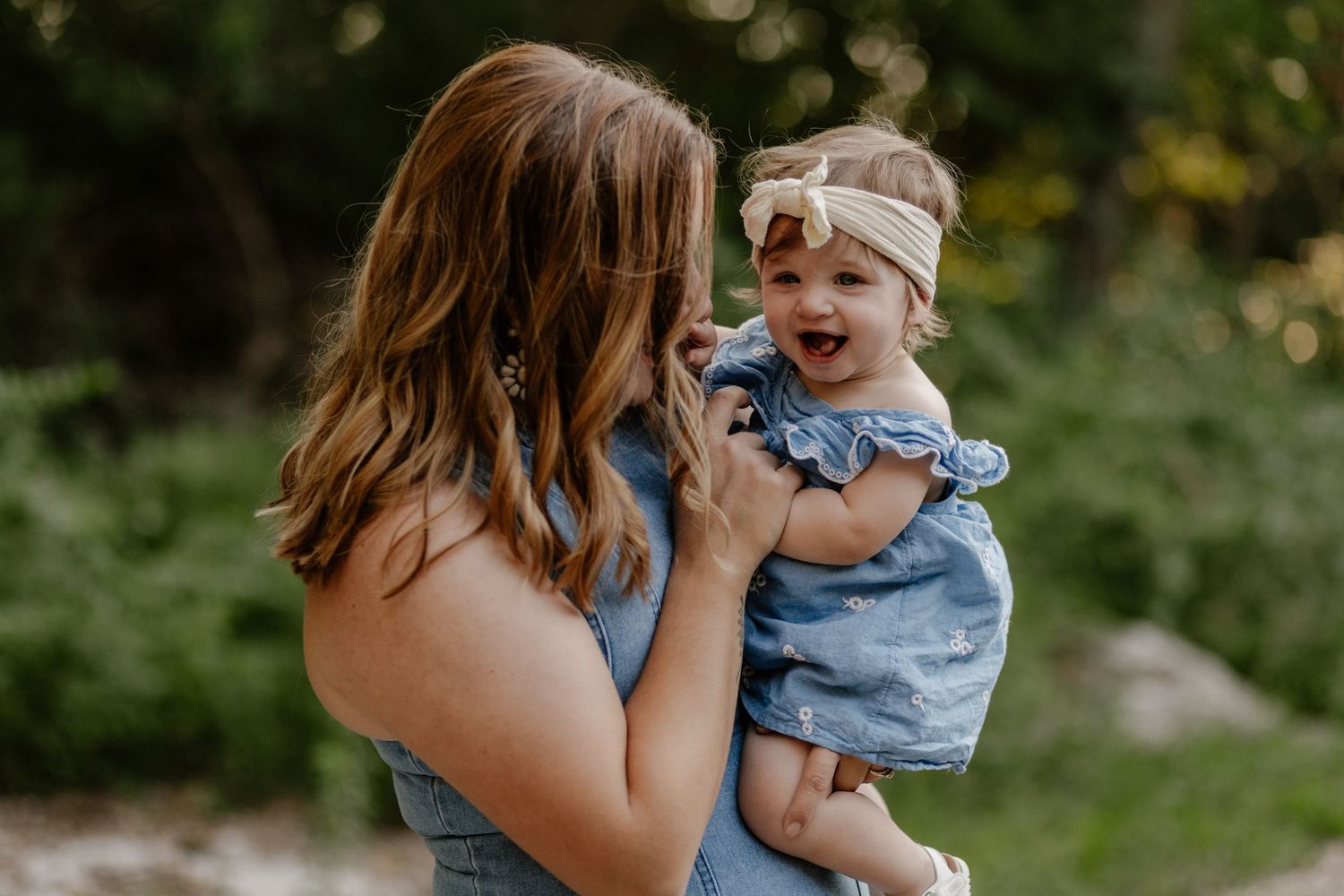 A mother in a blue dress holds her smiling baby girl wearing a denim outfit and bow headband outdoors.