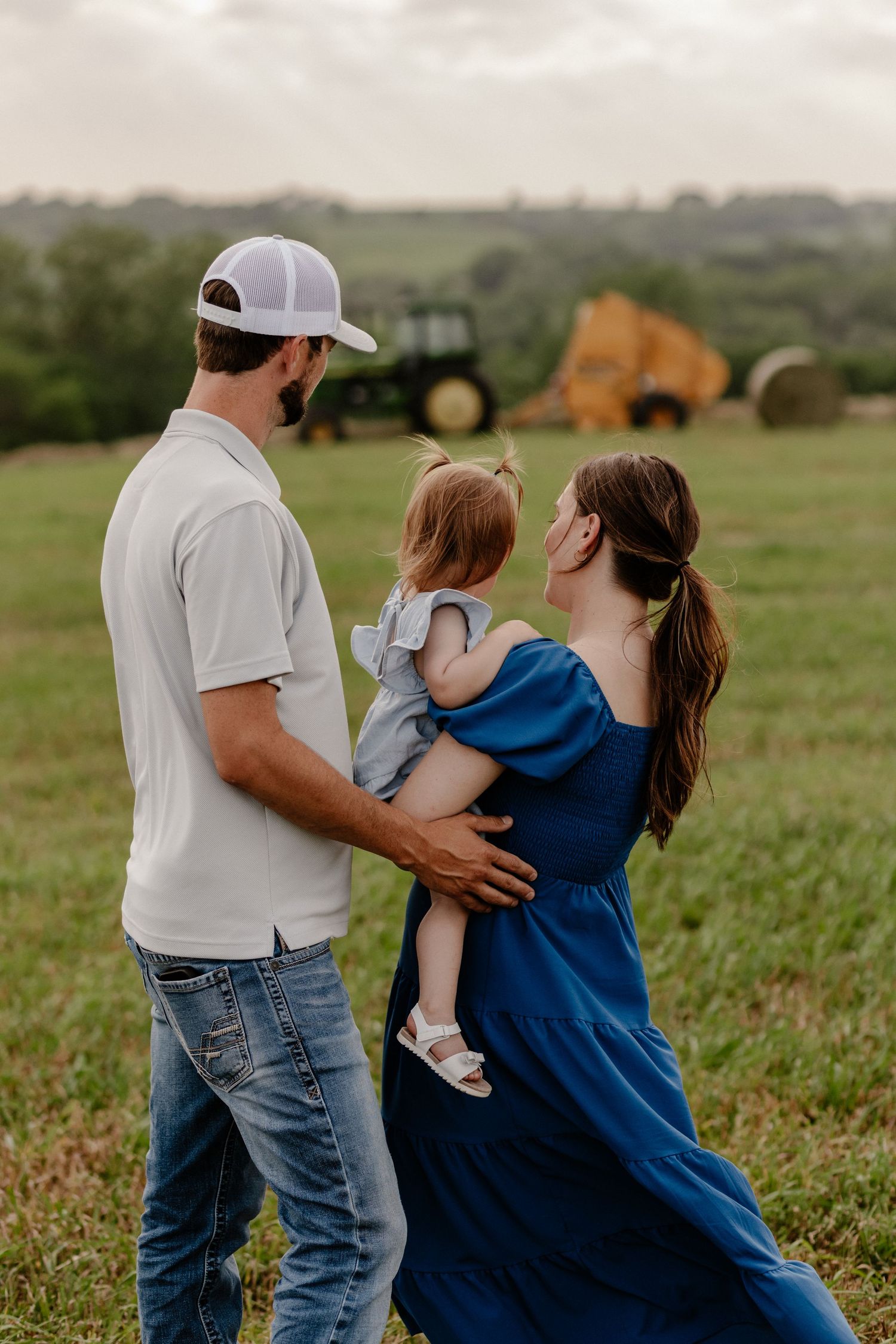 A young family with a toddler poses in a green field with a tractor and hay bale in the background.