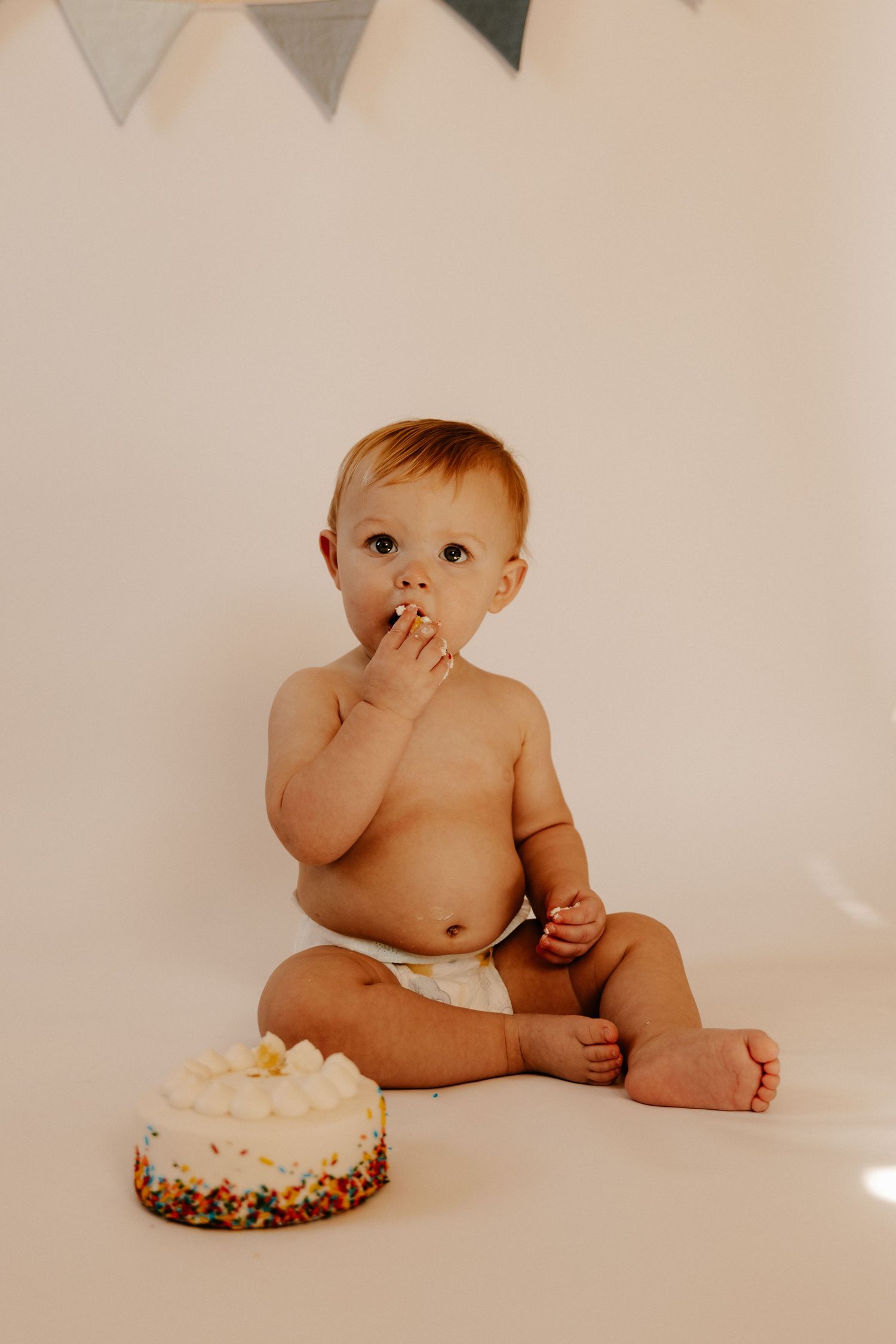 Baby in diaper sitting with birthday cake under grey bunting banner decoration.