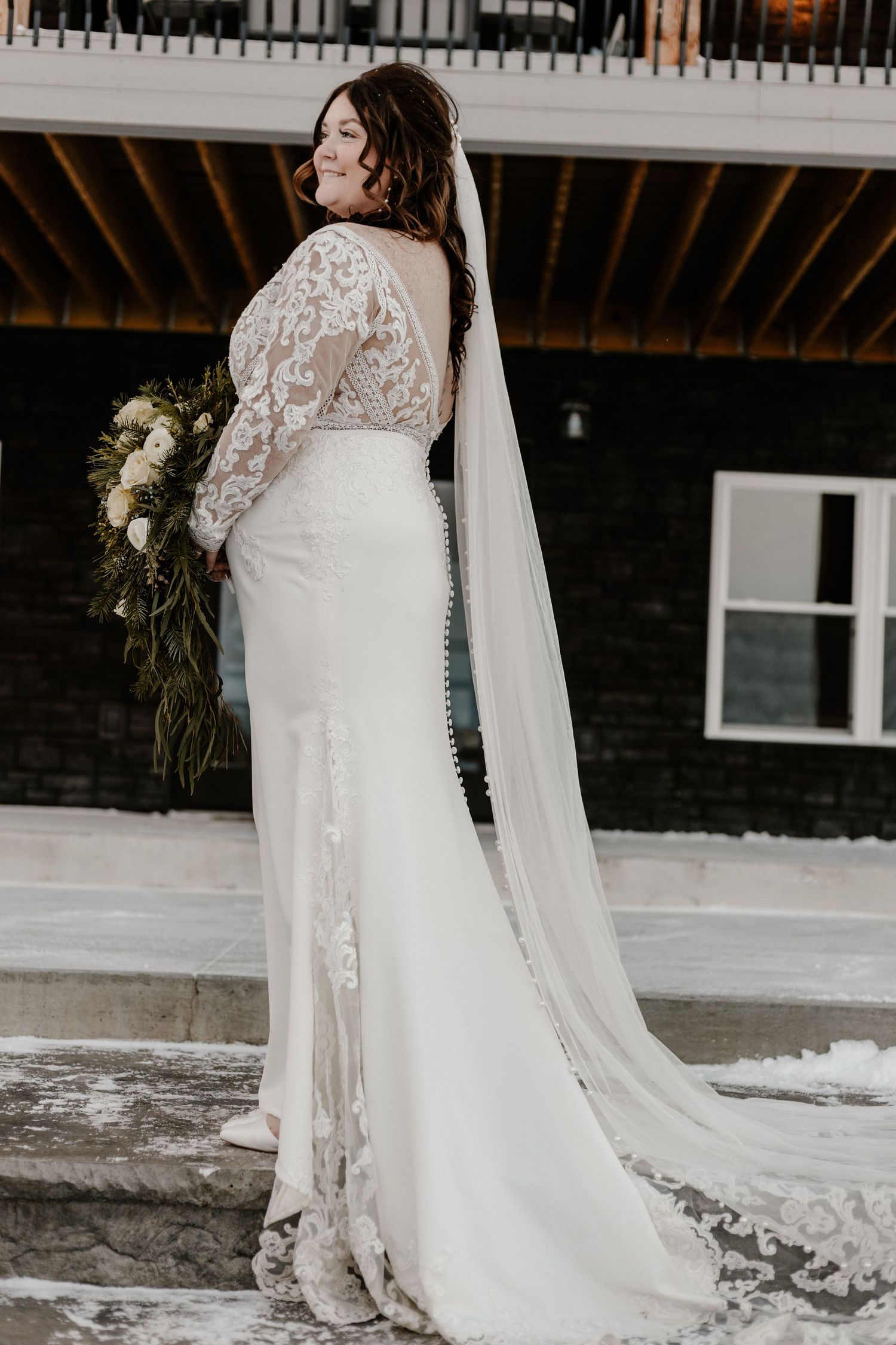 Elegant bride in lace long sleeve wedding gown with flowing veil poses outdoors in winter setting.