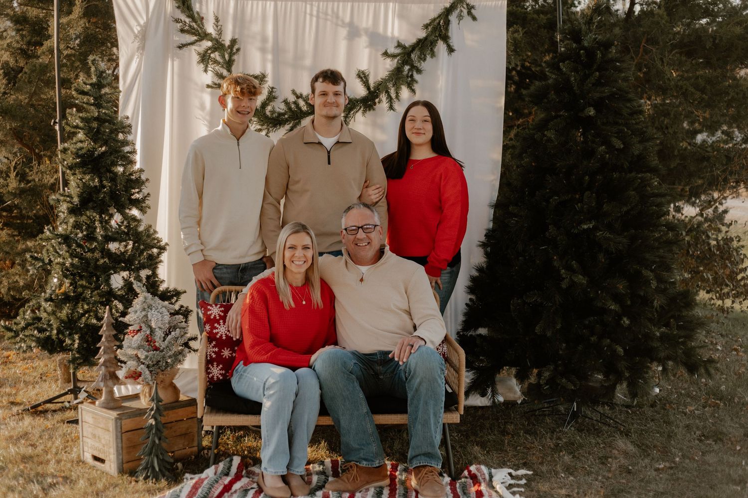 Holiday family portrait with five people arranged on a rustic bench against a white backdrop with greenery.