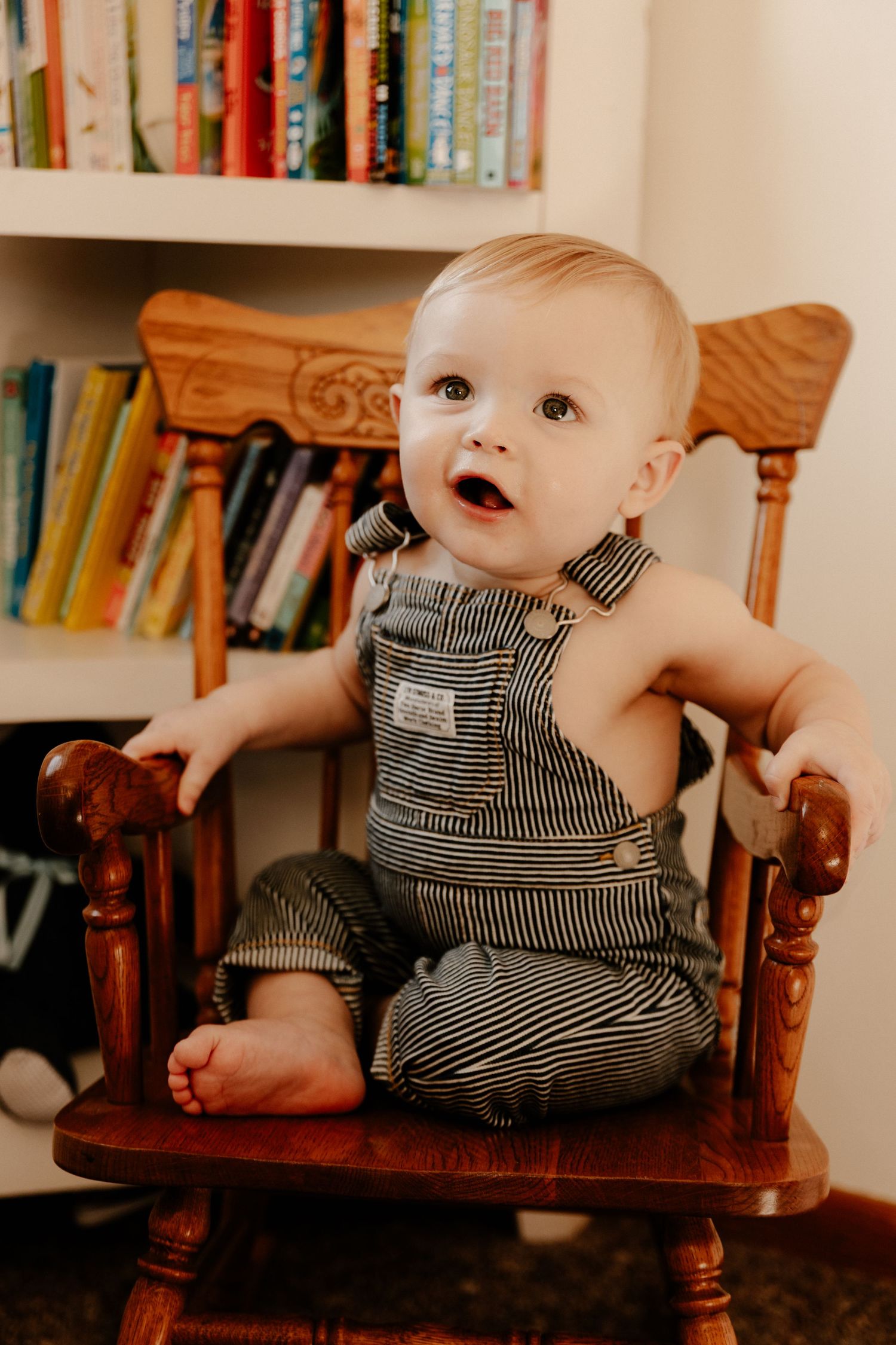 Baby in striped overalls sitting in rocking chair against bookshelf background.