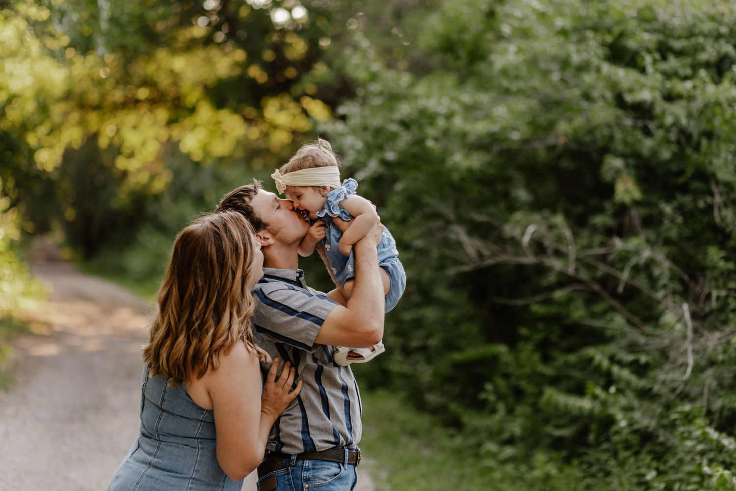 A couple kisses their baby outdoors on a sunlit path surrounded by lush green trees.