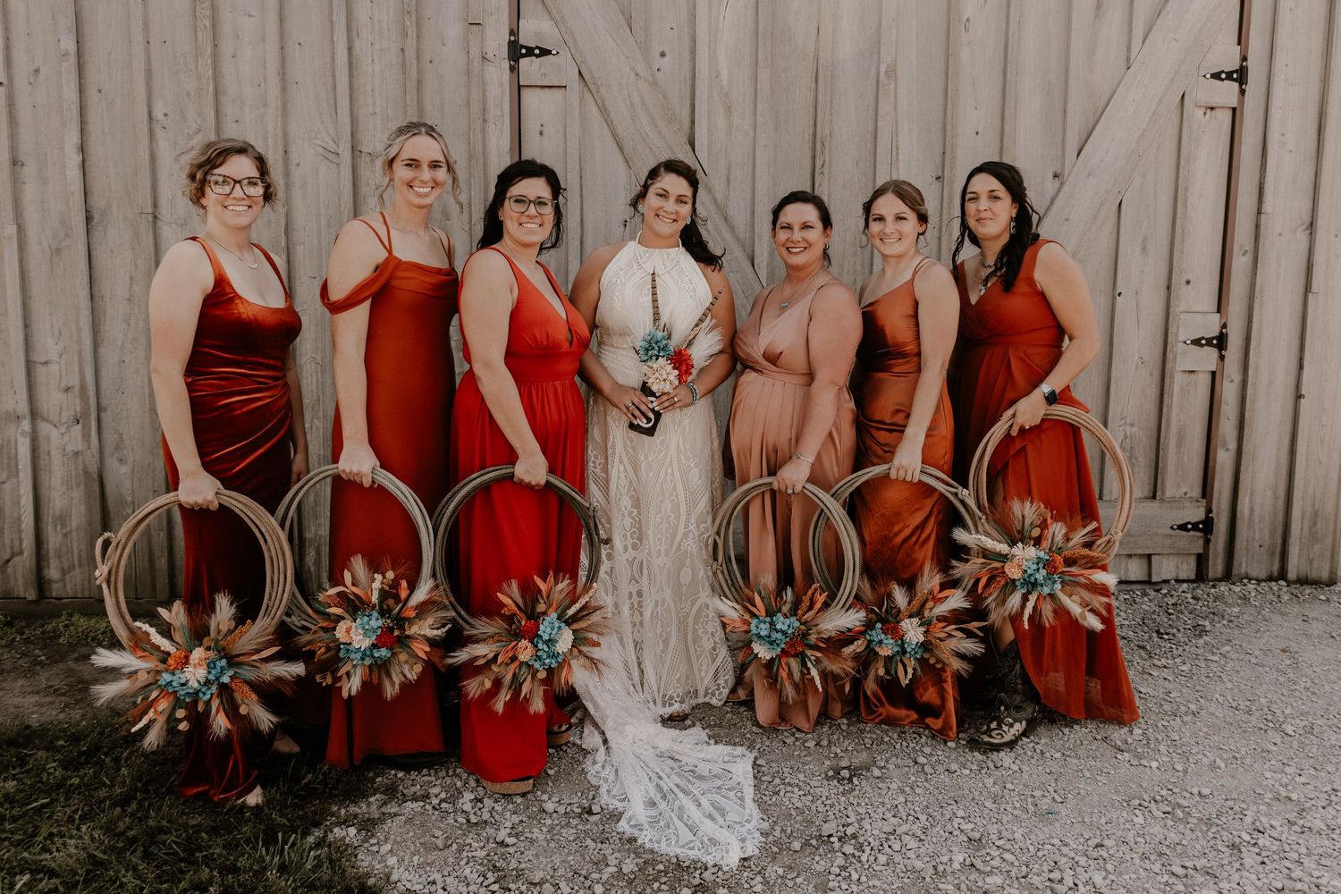 Wedding party in burgundy dresses poses against rustic white barn backdrop.