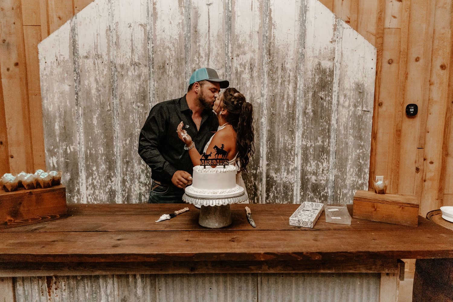Couple sharing an intimate moment with wedding cake on rustic wooden table against weathered backdrop.
