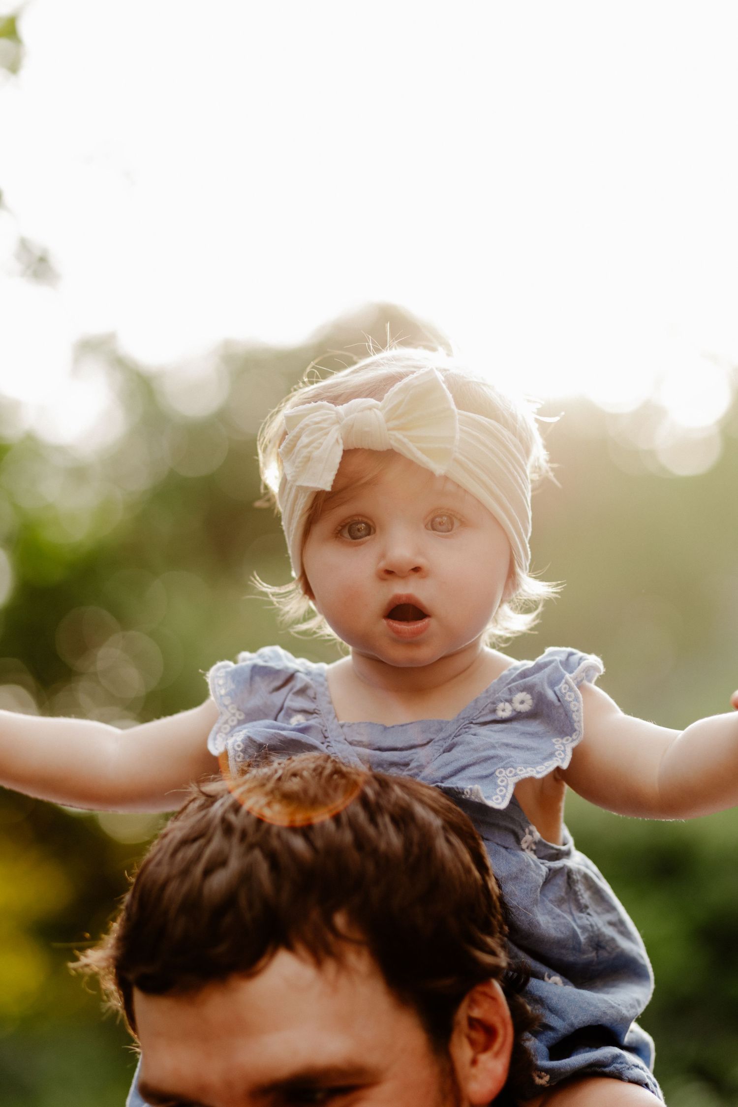 Adorable baby girl in blue dress and white bow headband sitting on father's shoulders outdoors in golden sunlight.