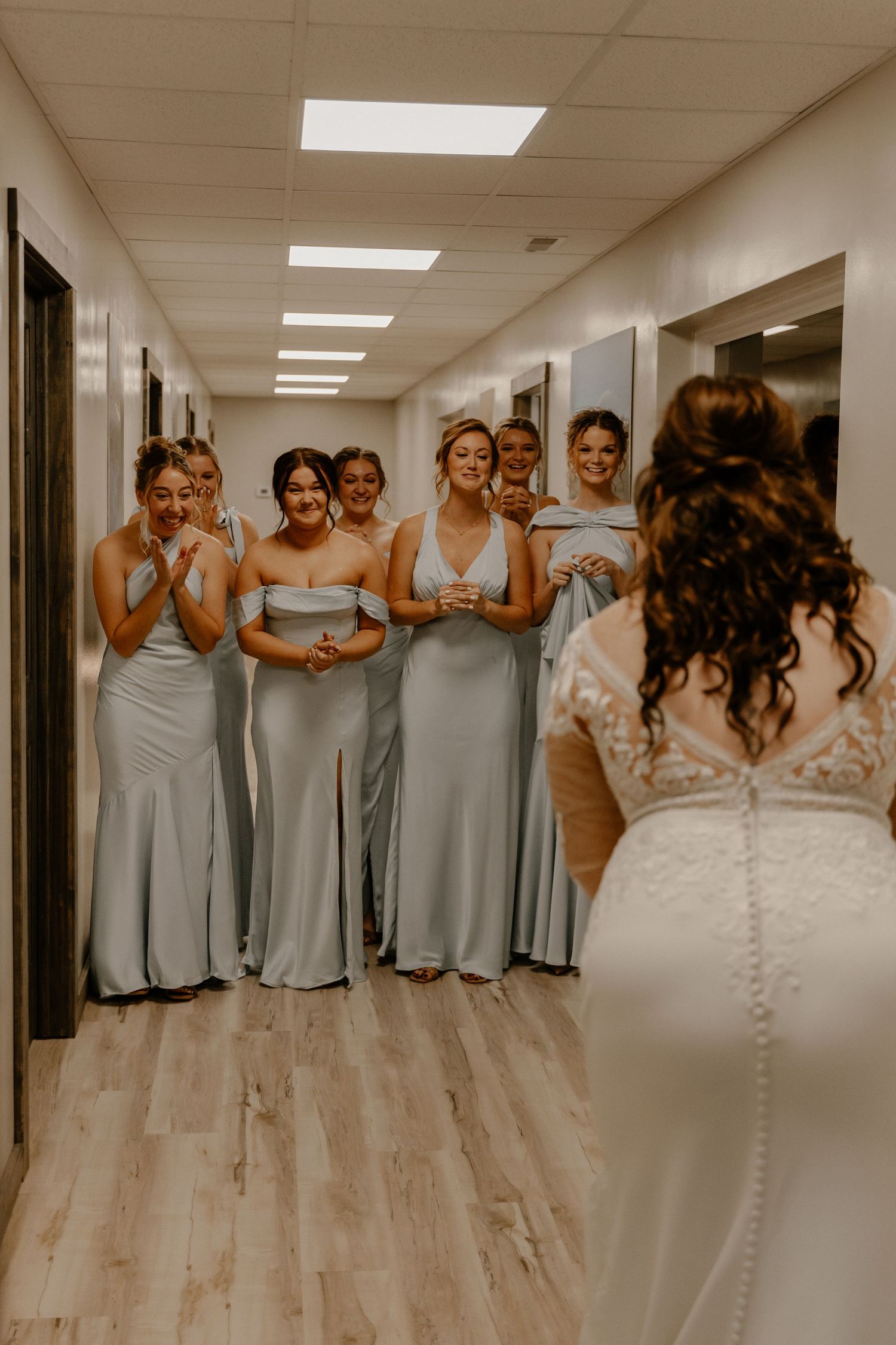 Bridesmaids in light blue dresses react to seeing the bride in a hallway.