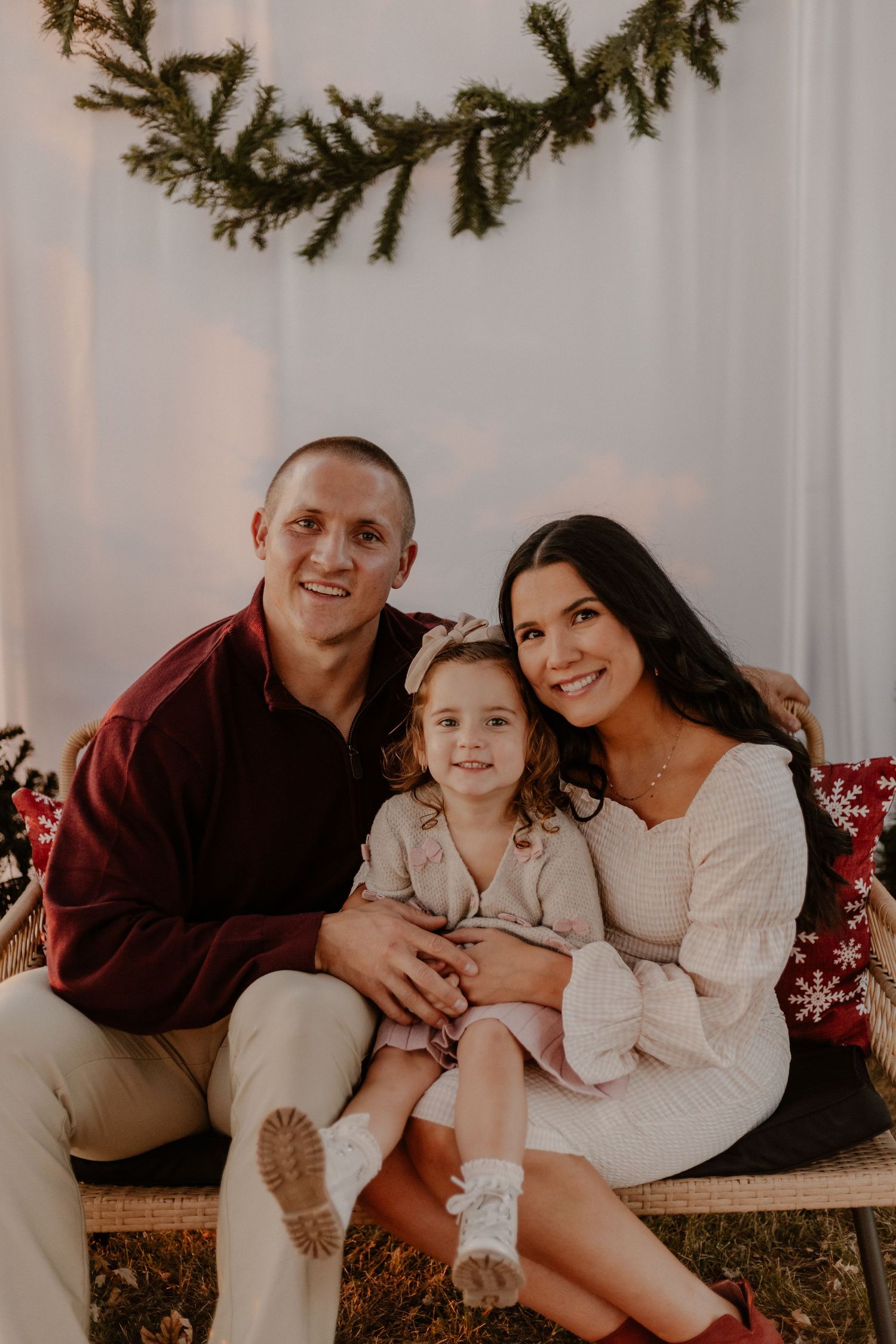 Holiday portrait session featuring a family of three sitting on a rustic bench with evergreen decor.