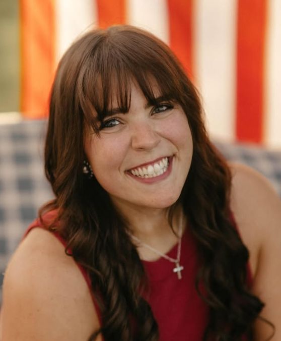 Smiling young woman with dark hair and bangs wearing a red top and cross necklace in front of an American flag.