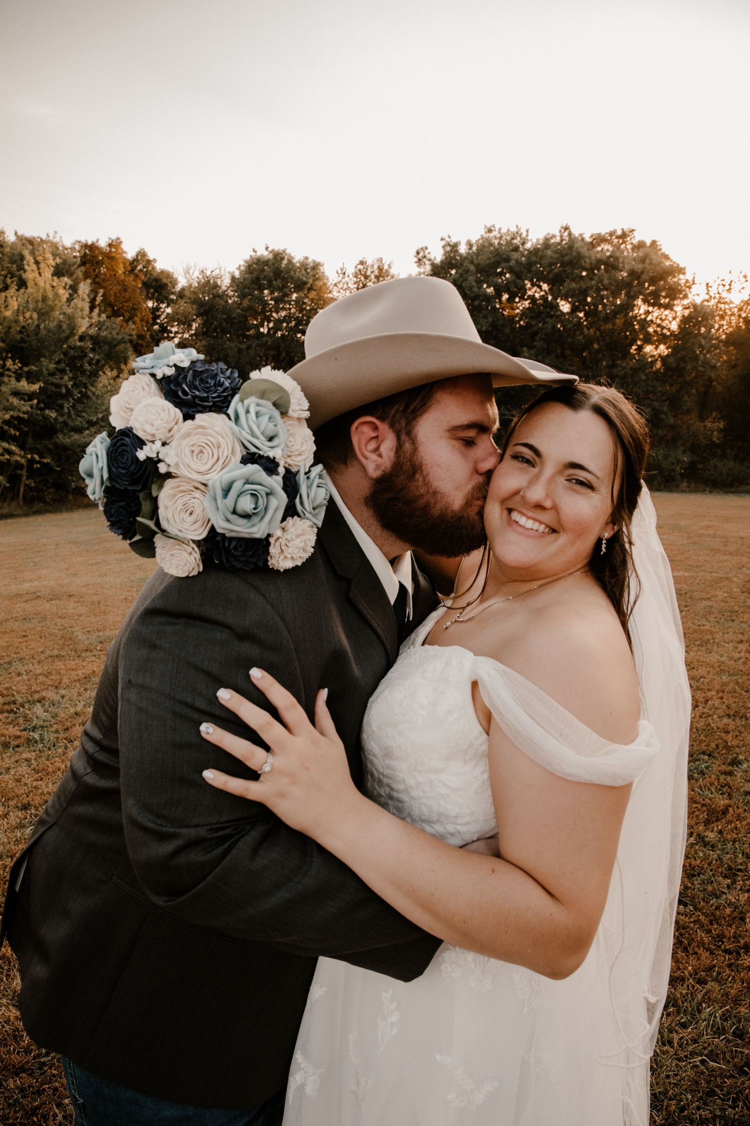 Newlyweds share romantic moment outdoors with blue and white rose bouquet at sunset.