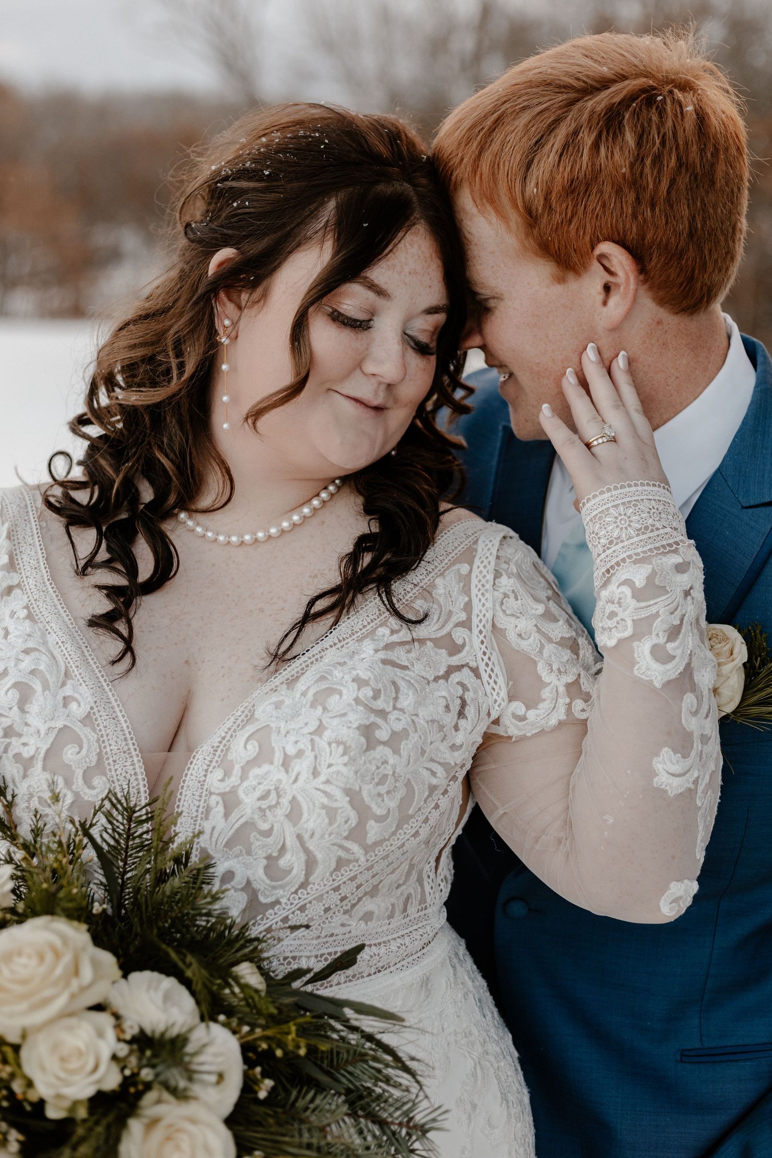 Romantic winter wedding portrait with couple in navy suit and lace dress against snowy backdrop.