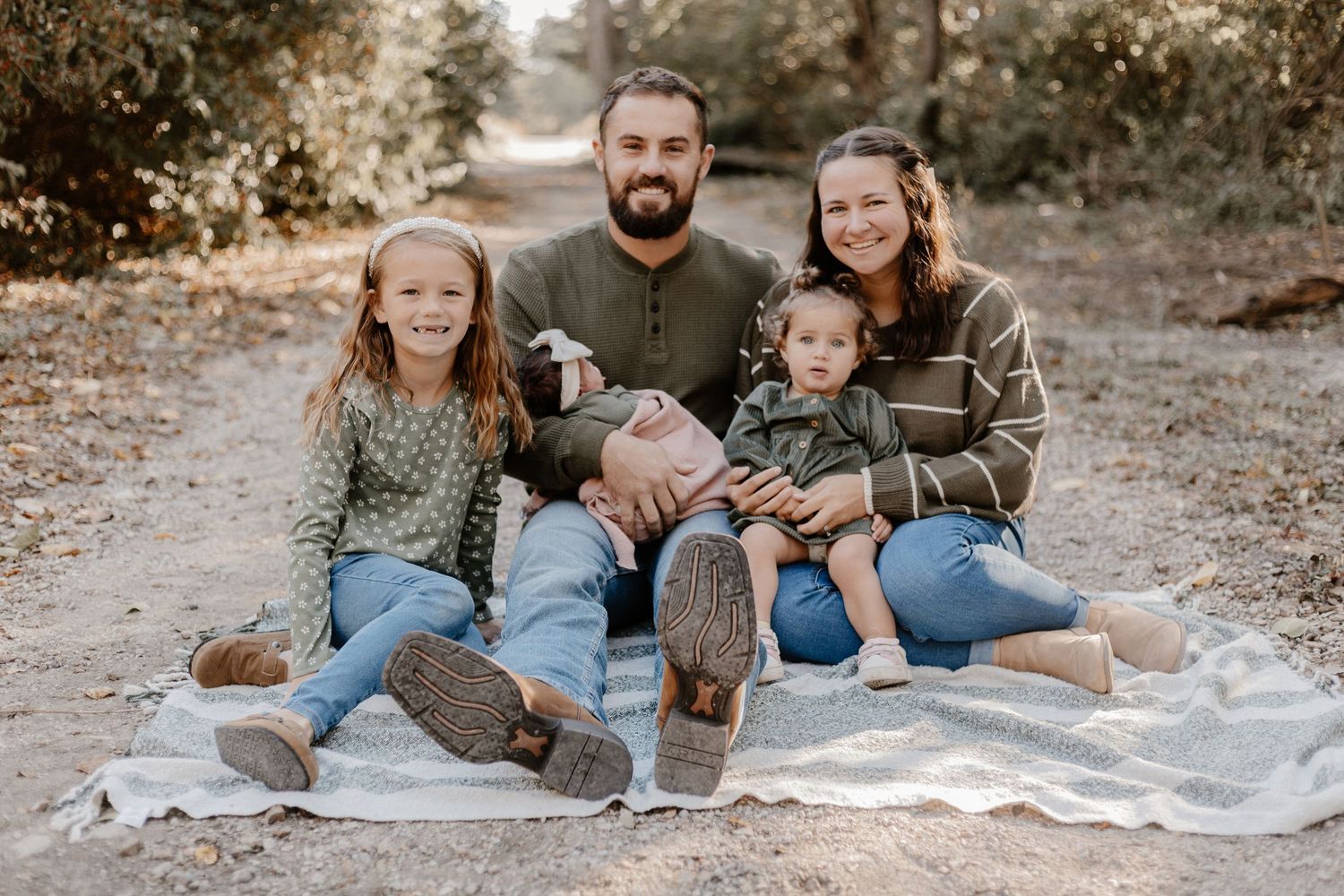 Family relaxing on white blanket during casual outdoor photo session in wooded setting.