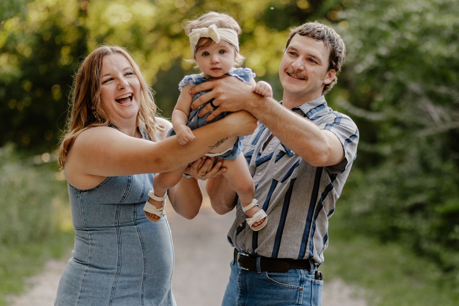Happy family with laughing parents lifting smiling baby outdoors on a sunny path surrounded by green trees.