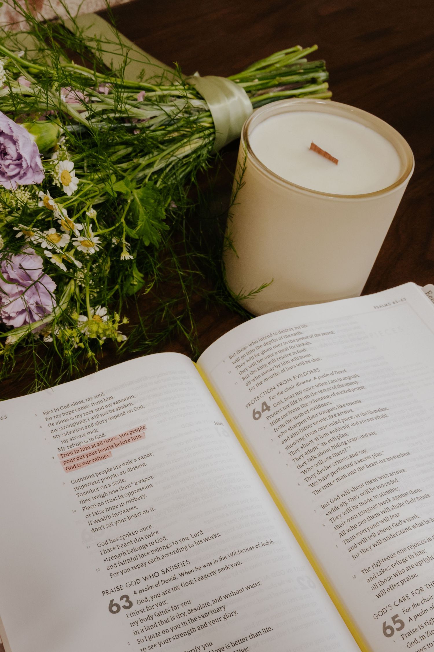 Open Bible beside a white candle and purple wildflower bouquet on a wooden table.