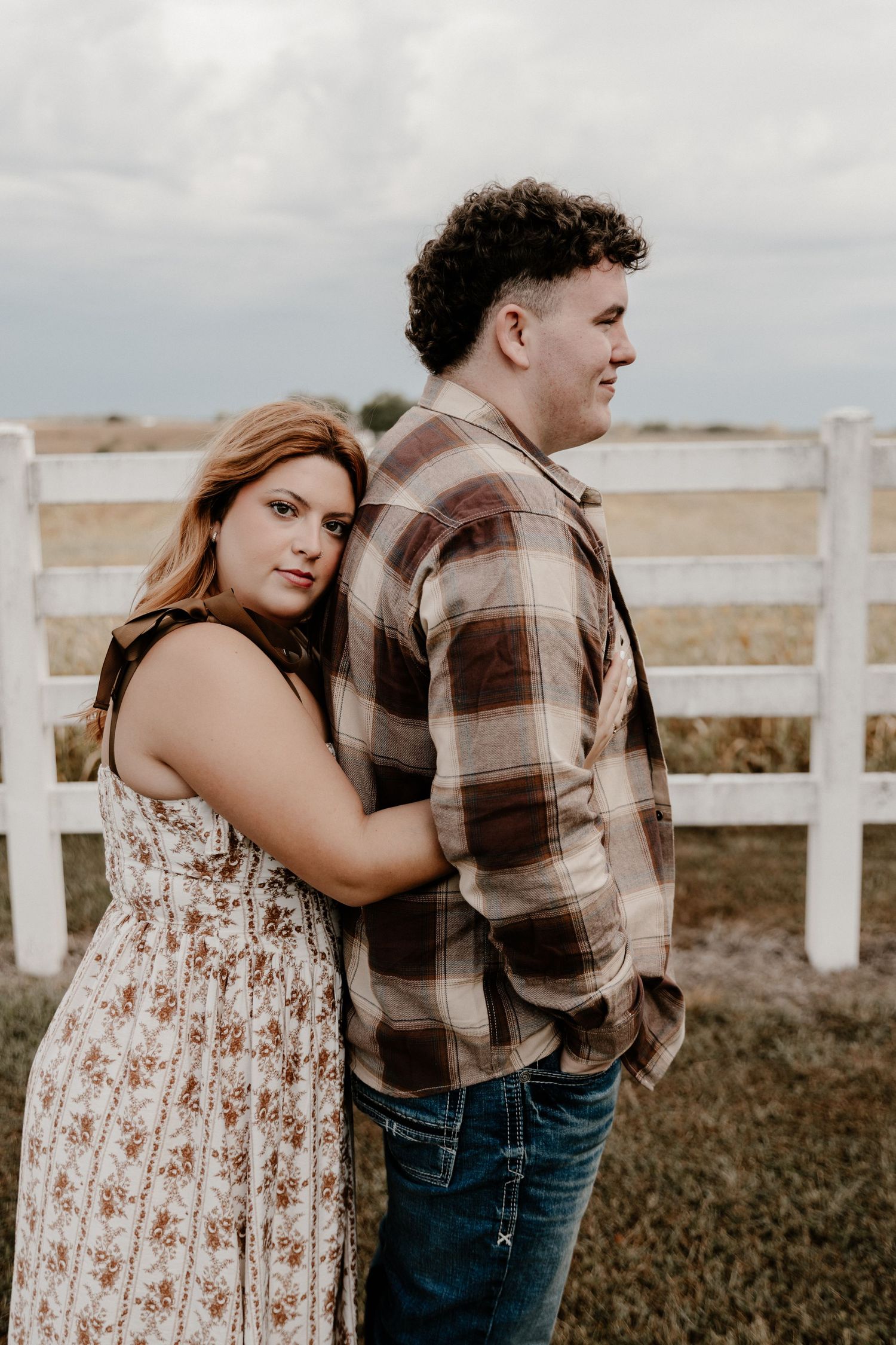 A couple poses near white fencing in a rustic countryside setting.