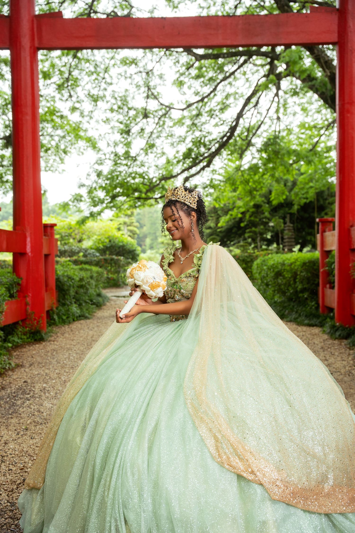 Woman in a sage green quinceañera ballgown with gold crown holds a white floral bouquet beneath a red torii gate in a lush garden.