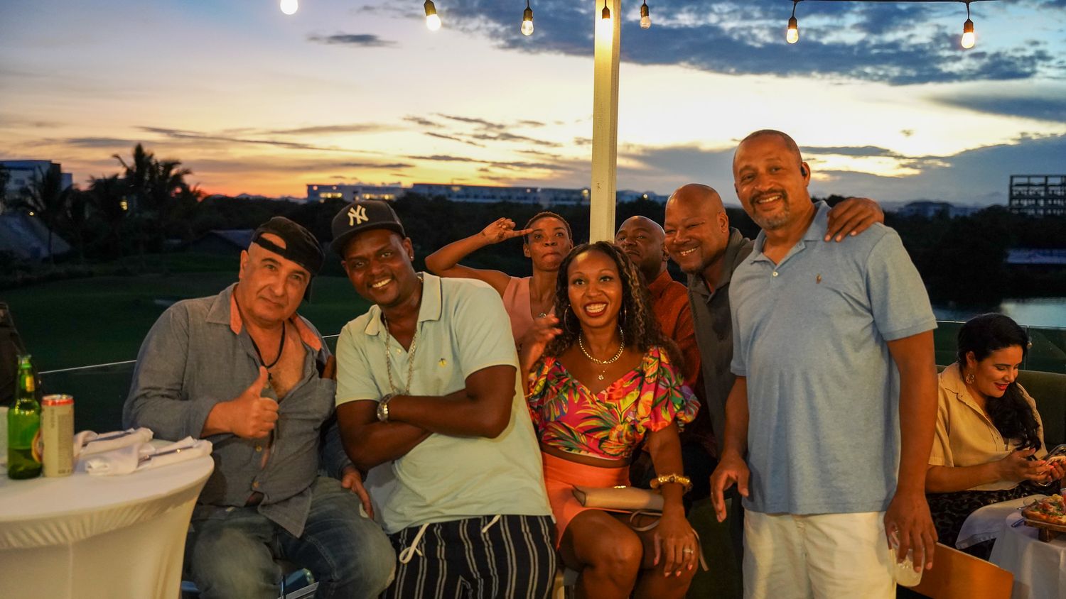 A group of smiling friends poses close together at an outdoor sunset gathering with string lights, drinks and a tropical view.