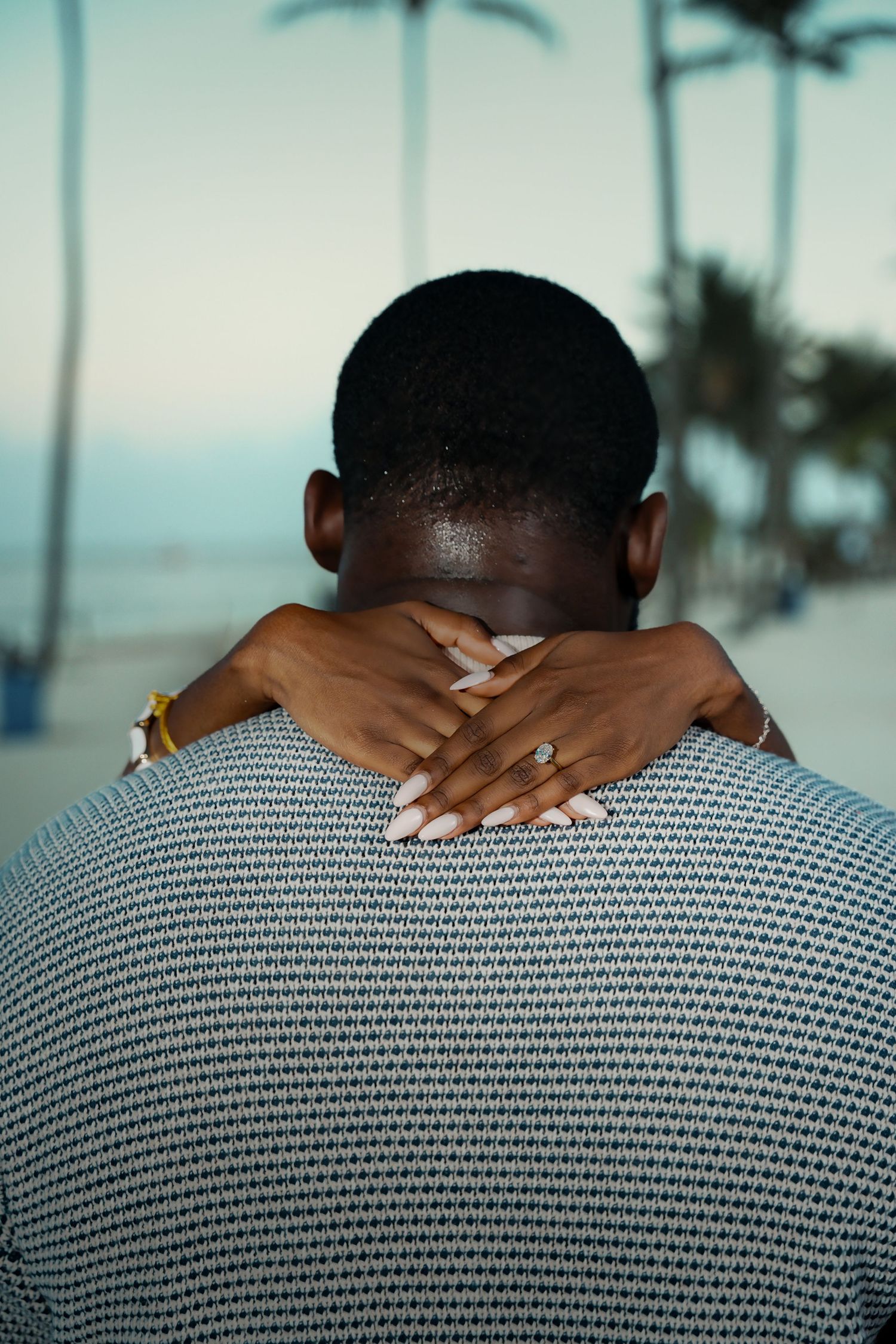 Woman's arms wrapped around a man's neck, wearing a ring and bracelet, in a romantic embrace on a tropical beach with palm trees.