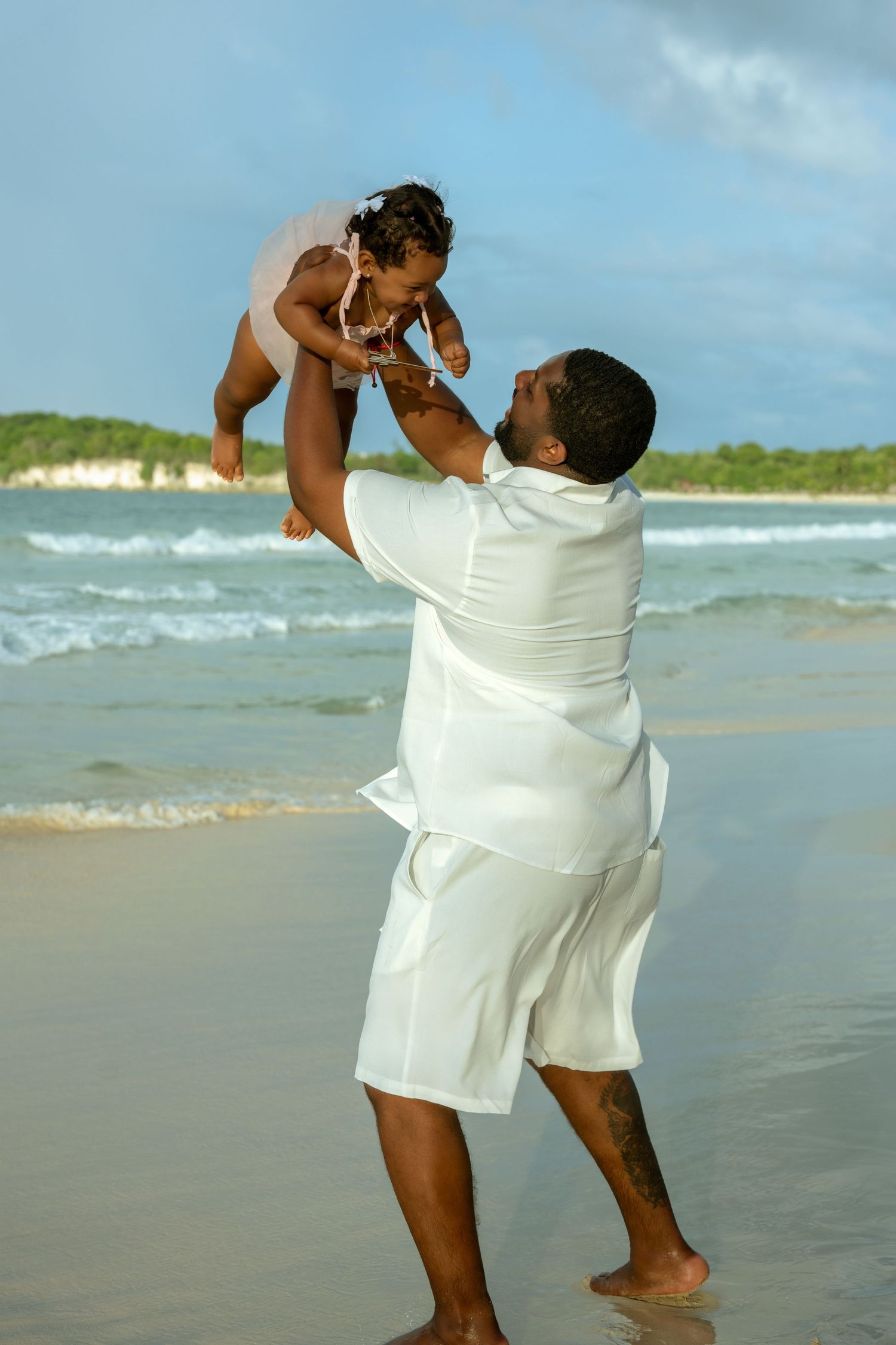 Man in white linen clothes lifts a laughing baby into the air while standing barefoot on a sunny tropical beach by the ocean.