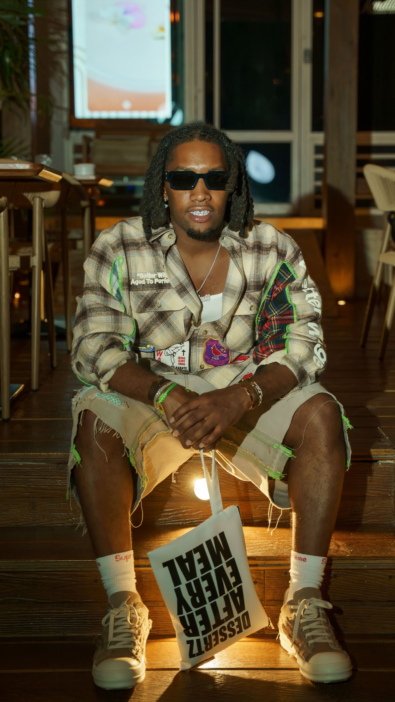 Man in dark sunglasses and a plaid shirt with patches sits on wooden steps holding a white tote bag reading Dessert After Every Meal.