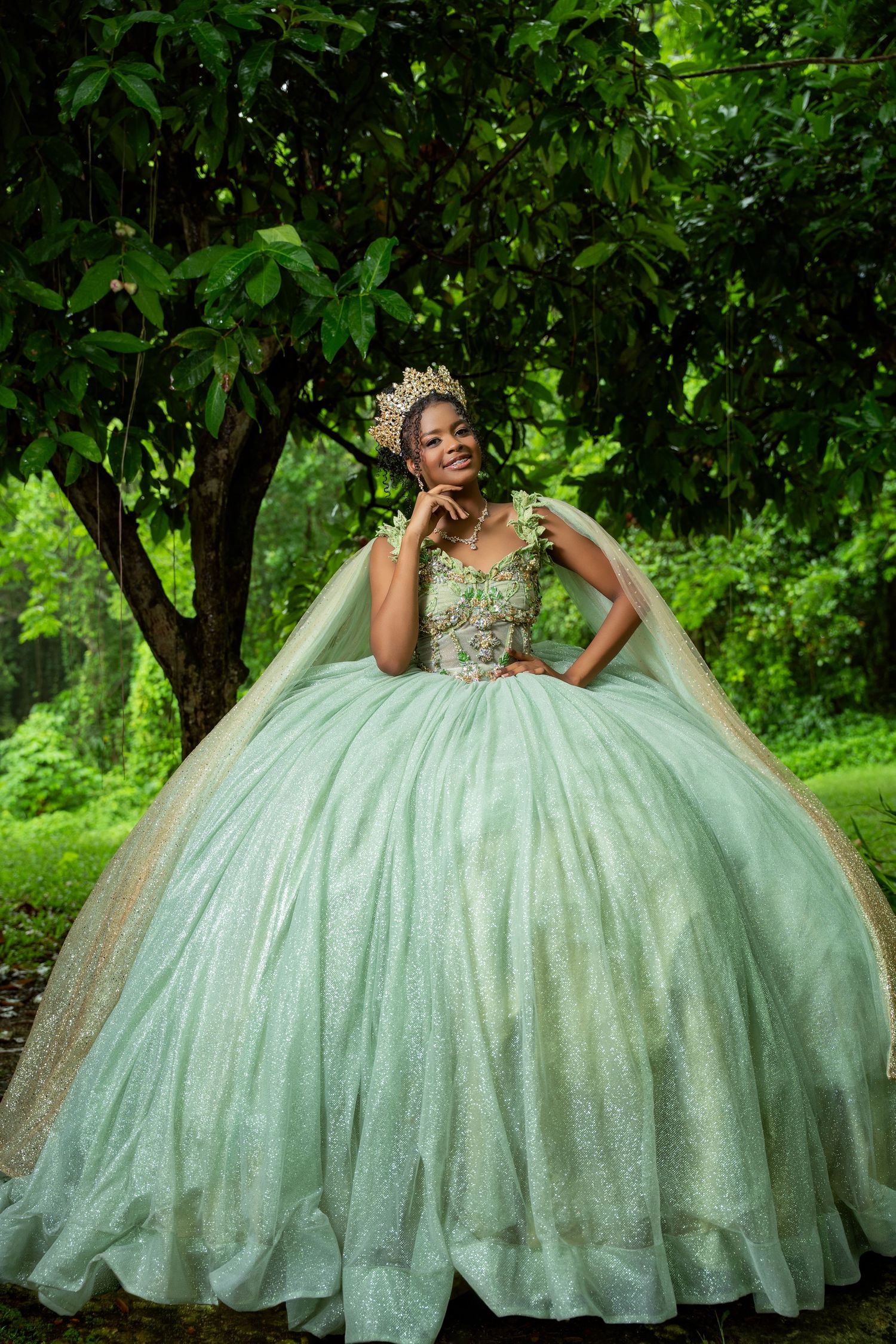 Woman in a sparkling mint green quinceañera ball gown with a cape and gold crown posing beneath lush green trees.