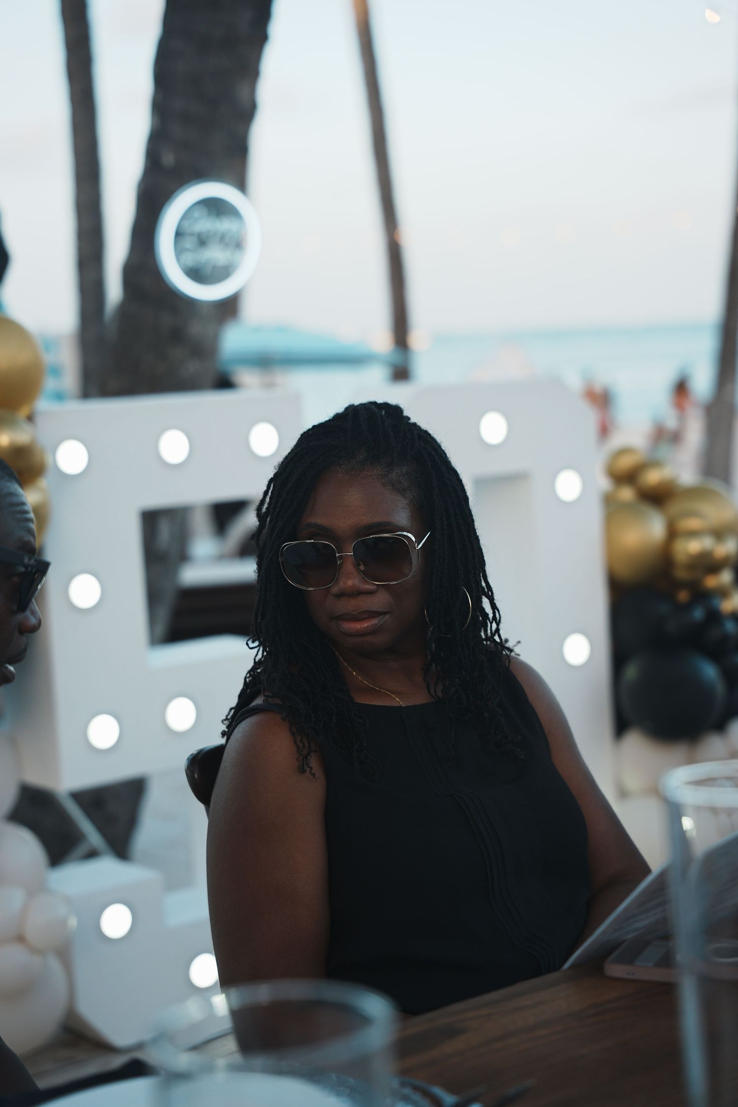 Woman in sunglasses and a black sleeveless top sits at a table at a beachfront party with lit marquee letters and gold and black balloons.
