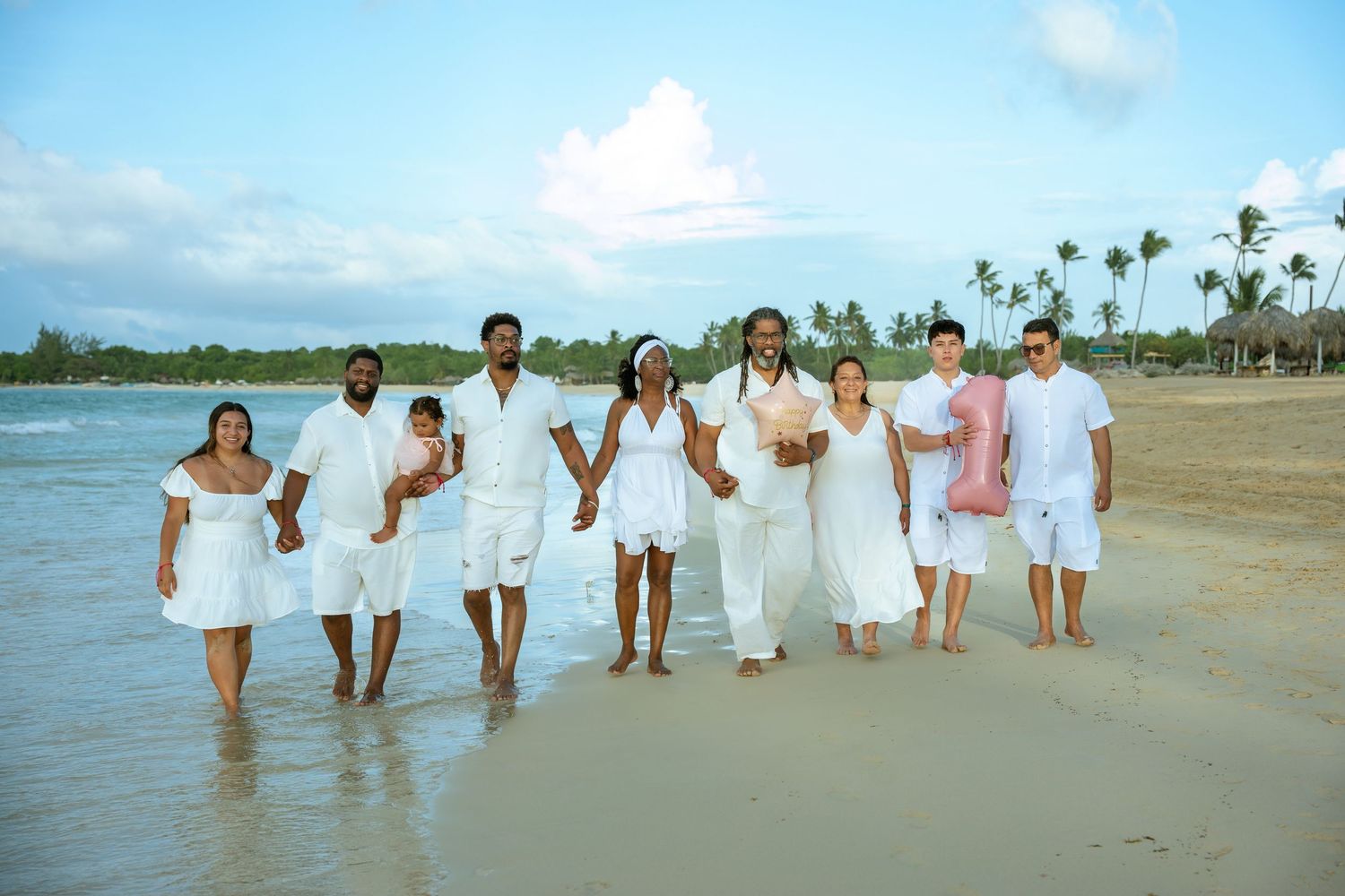 Family in white outfits walking hand in hand along the shoreline of a tropical beach with palm trees under a blue sky.
