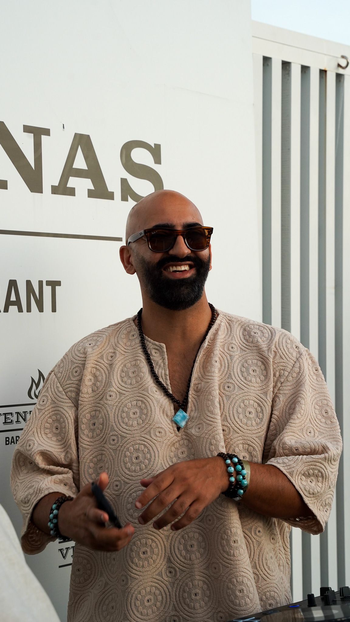 Smiling bald man with a beard, sunglasses and beaded bracelets wears a beige embroidered tunic and turquoise pendant outside a white restaur