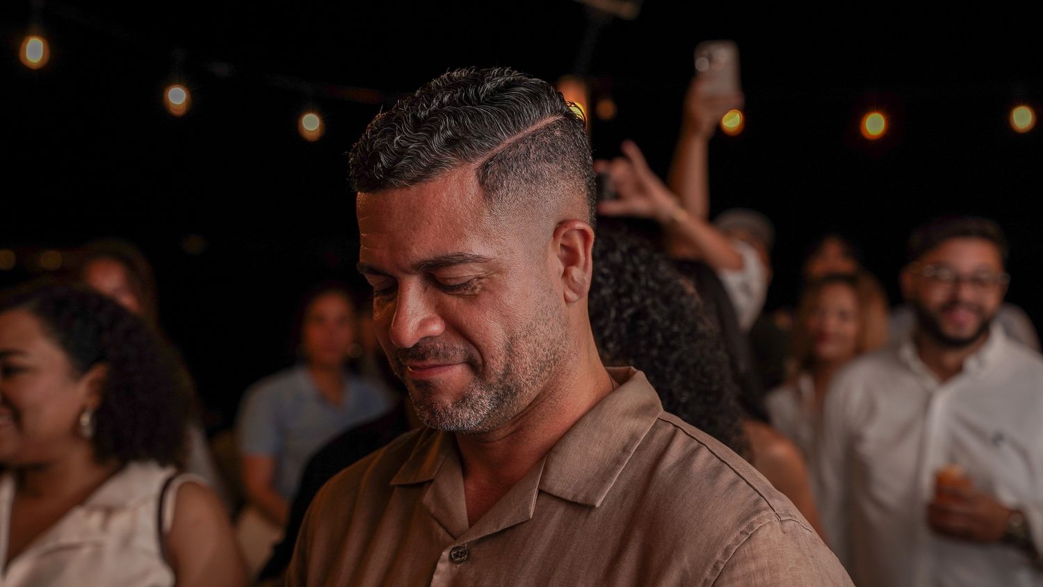 Smiling man with short styled hair and a beard in a tan button-up shirt at a lively evening party with string lights and guests behind him.