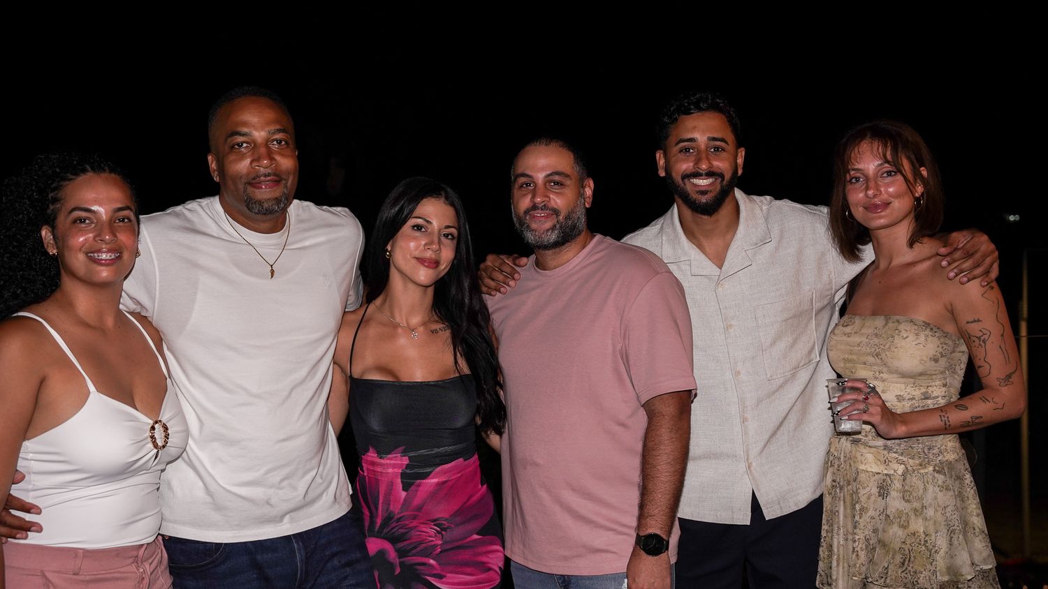 Six smiling friends in casual summer attire pose arm in arm at a nighttime outdoor gathering against a dark background.