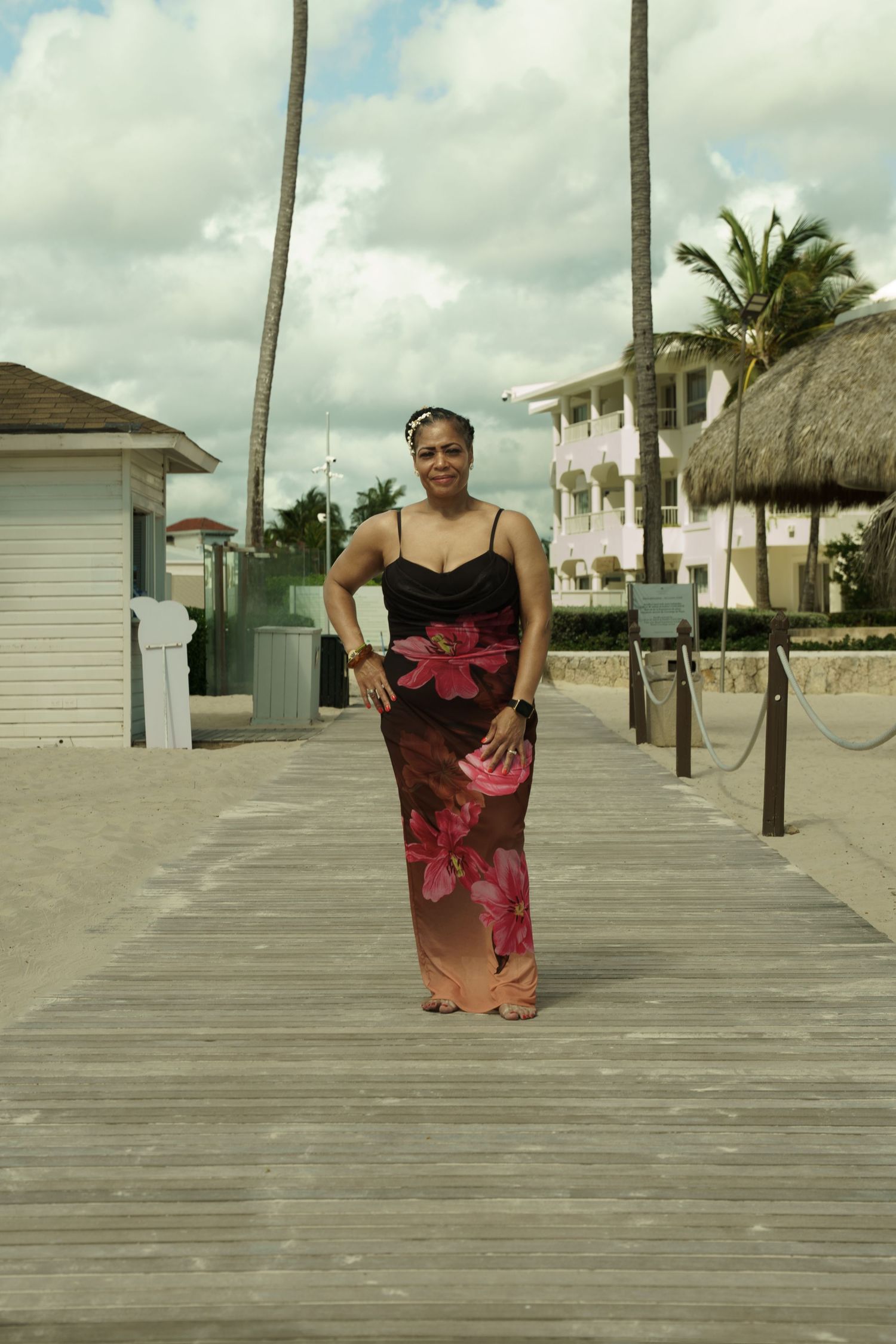 Woman in a long floral maxi dress poses on a wooden beach boardwalk near palm trees, a thatched hut, and a resort building.