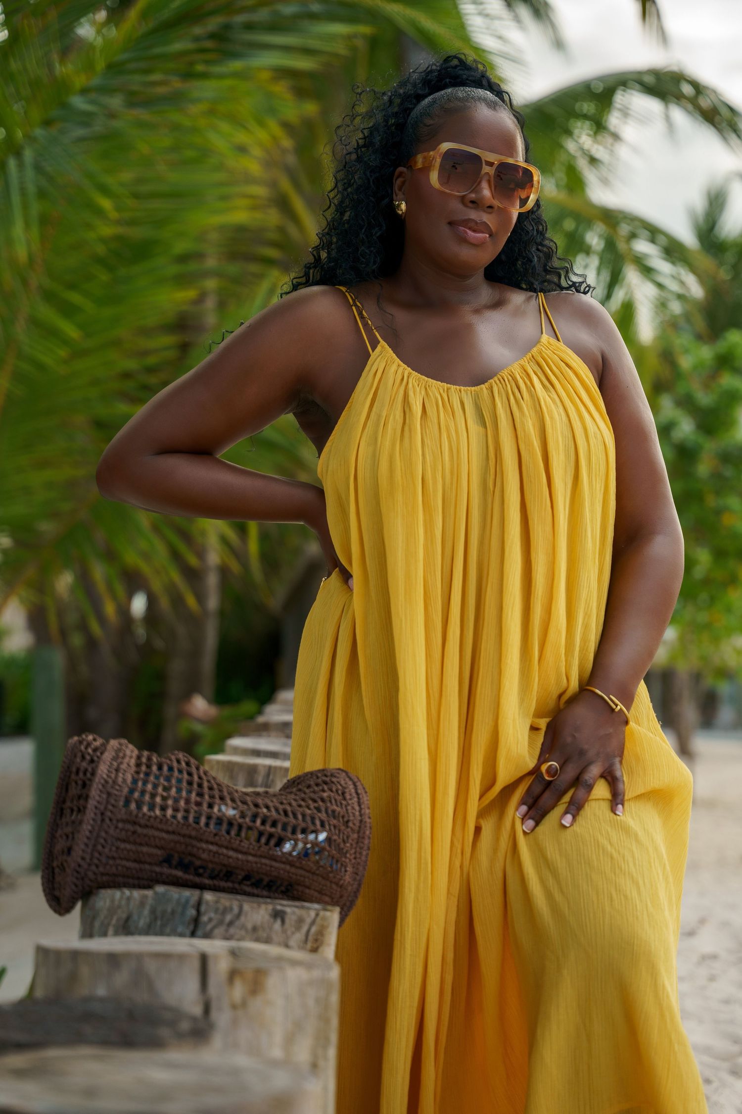 Woman in a flowing yellow sleeveless maxi dress and tinted sunglasses poses beside a woven basket bag near palm trees on a tropical beach.