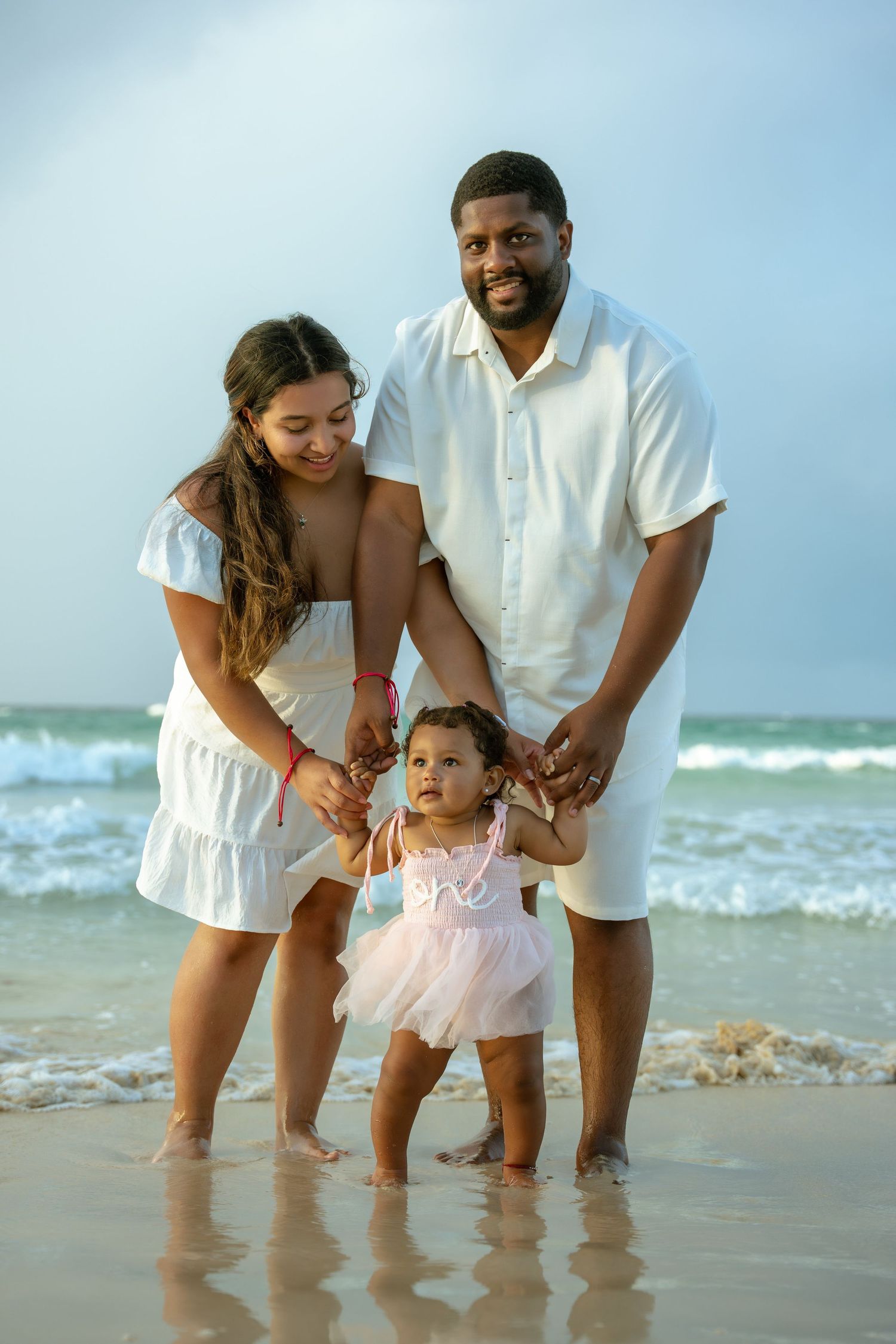 Smiling couple in white outfits hold hands with their toddler in a pink tutu dress as she stands on the wet sand by the ocean.