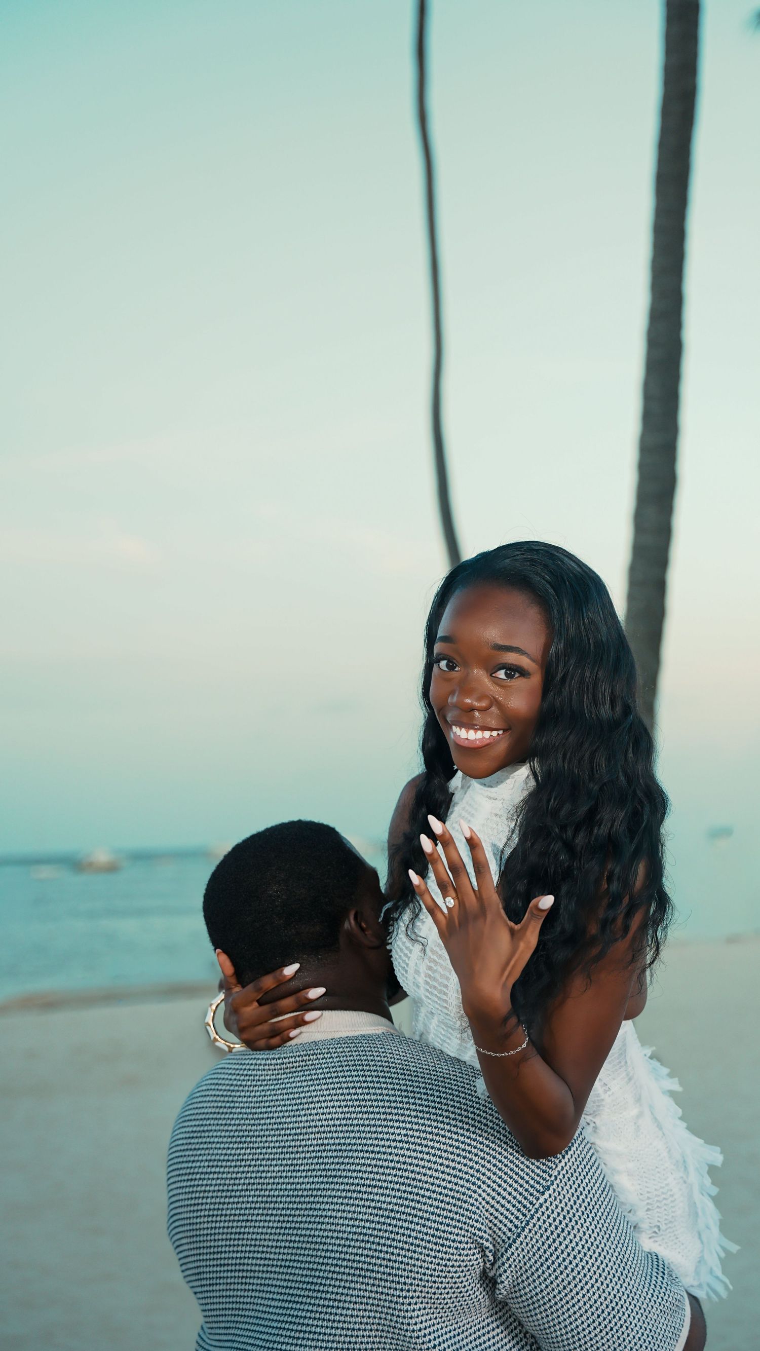 Smiling woman in a white dress shows her ring on a beachside engagement as a man in a gray sweater faces her, palm trees and ocean behind.