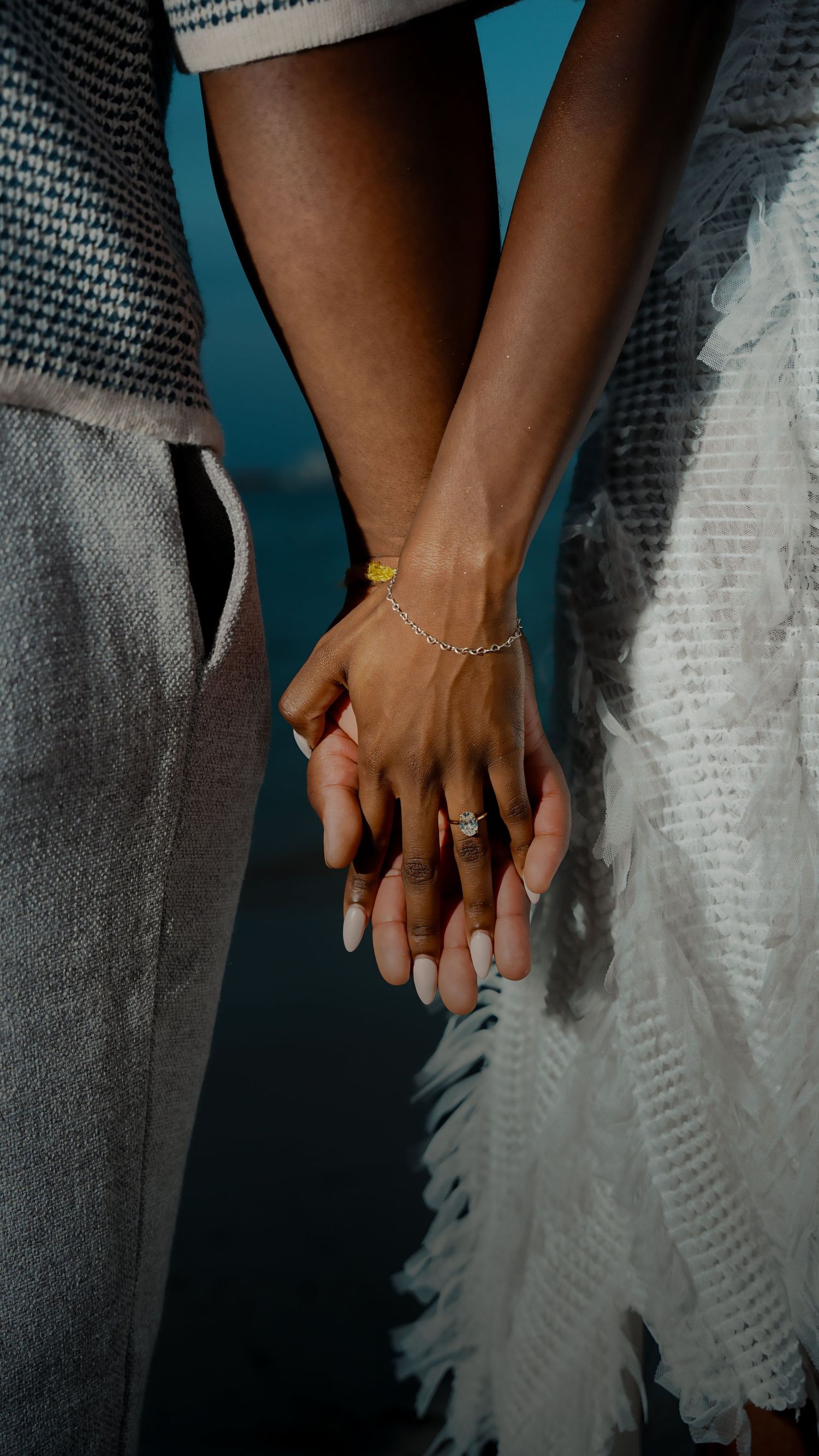 Close-up of two people holding hands at dusk, wearing a gold bracelet and a ring, with textured gray and white feathered clothing.