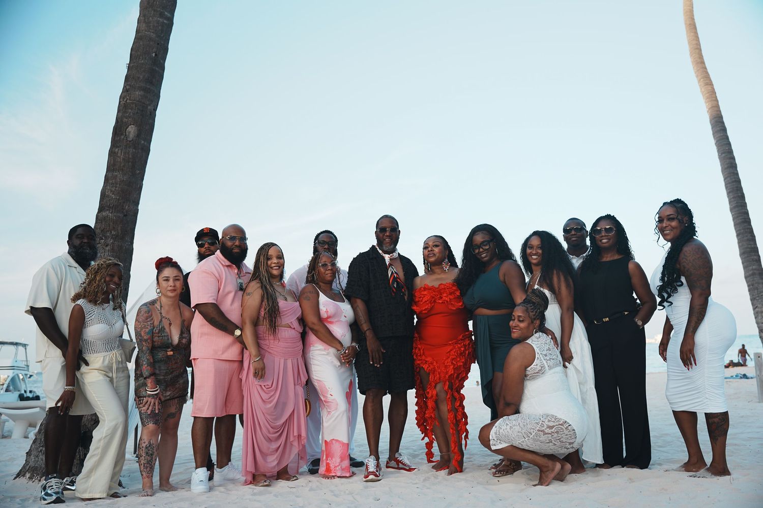 Group of smiling friends in colorful summer outfits posing together on a sandy tropical beach near palm trees at sunset.