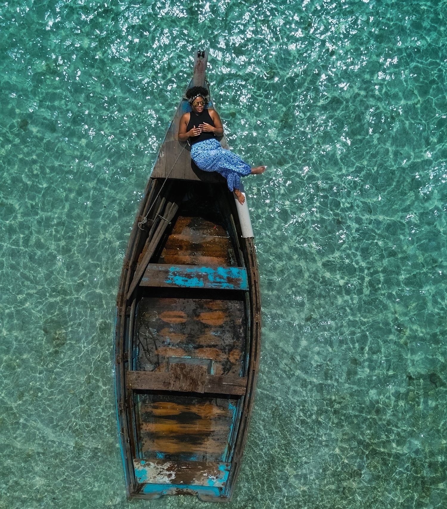 Aerial view of a woman in a blue patterned dress sitting at the bow of a weathered wooden boat on clear turquoise tropical water.