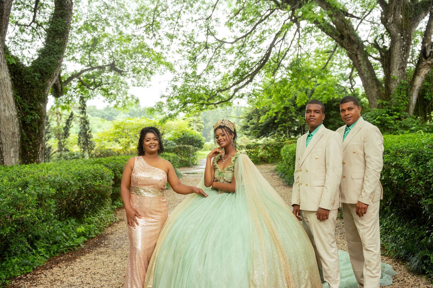 Quinceañera in a mint green ball gown poses outdoors with a woman in a rose gold dress and two men in beige suits under lush green trees.