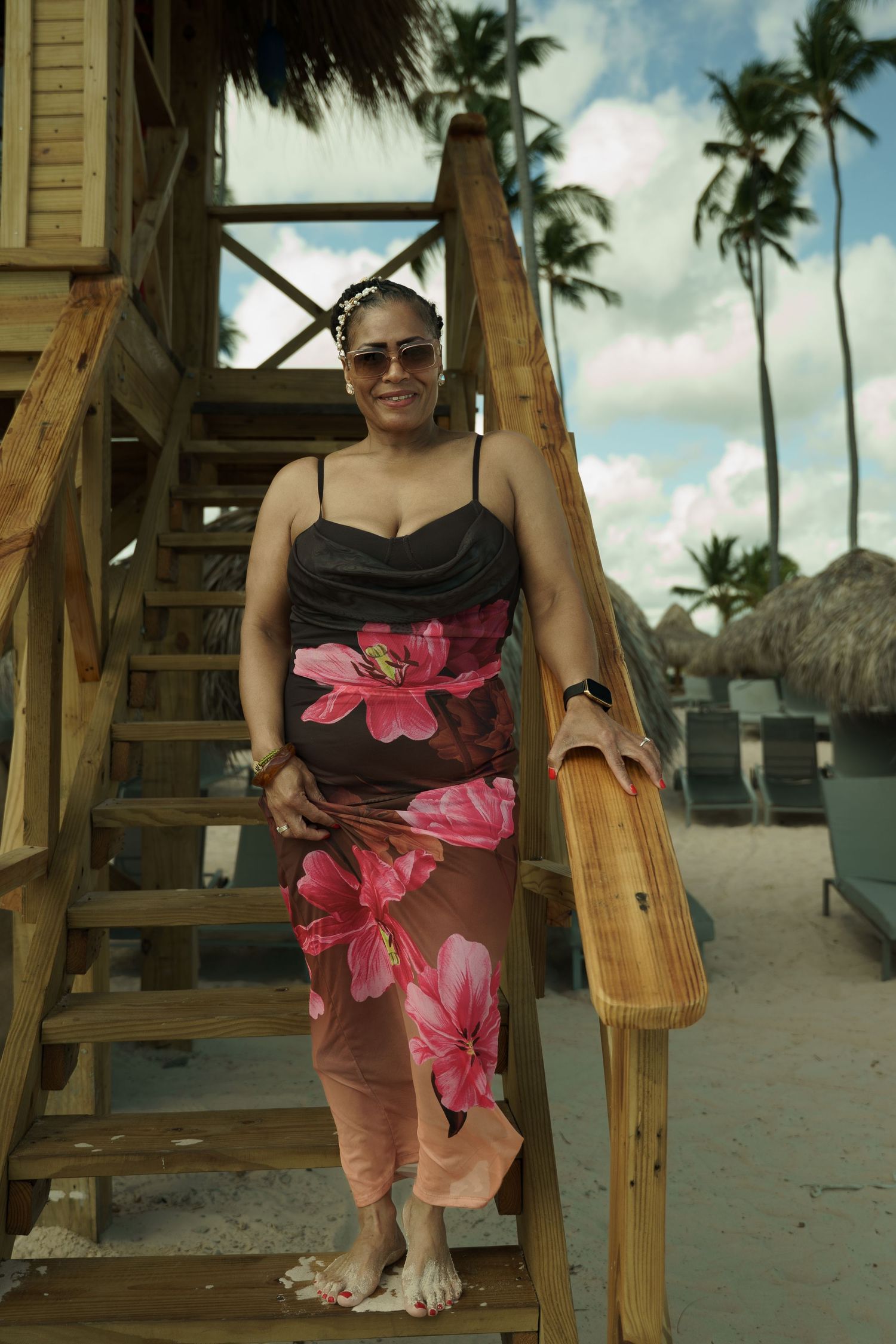 Woman in sunglasses and a pink floral sarong dress stands barefoot on wooden beach stairs near palm trees and thatched huts.