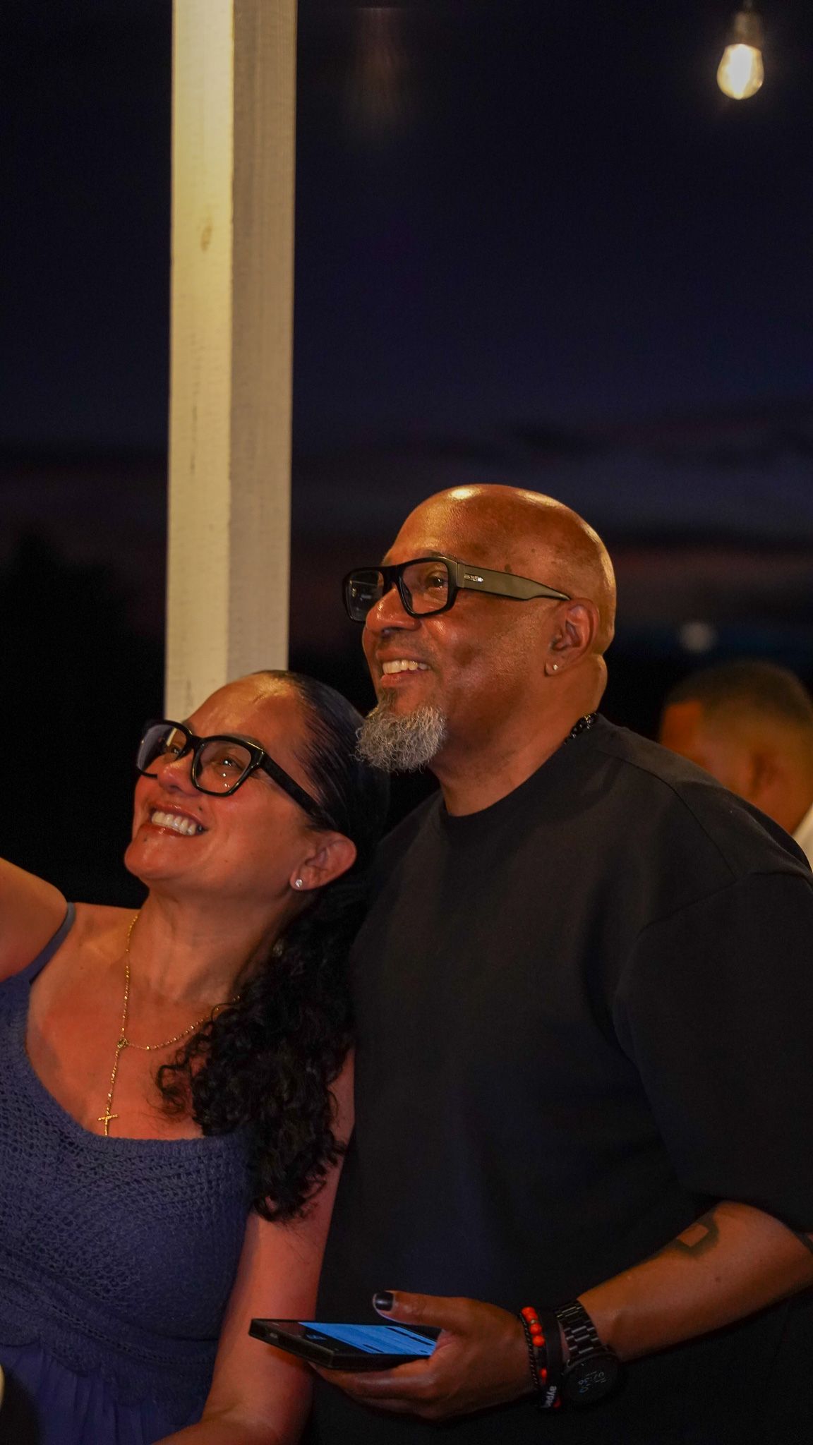 Smiling man and woman in glasses look up together at an evening outdoor gathering, with string lights and a dusky sky behind them.