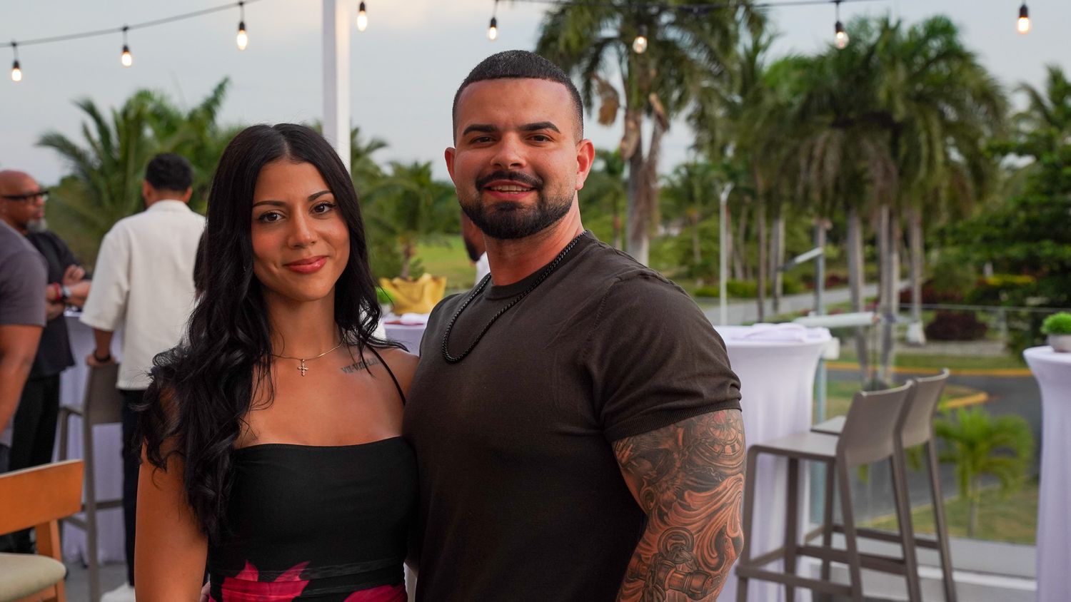 Smiling couple posing together at an outdoor tropical resort terrace with palm trees and string lights at sunset.
