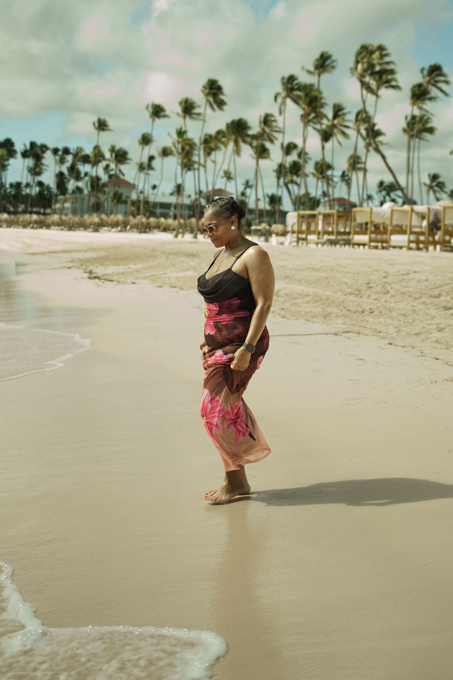 Woman in a black top and pink floral sarong walking barefoot along a sandy tropical beach with palm trees and cloudy sky behind her.