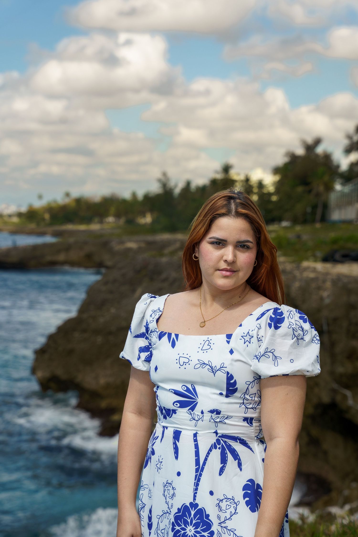 Woman with auburn hair wearing a blue and white floral puff-sleeve dress poses on a rocky coastal cliff beside the ocean under a cloudy sky.