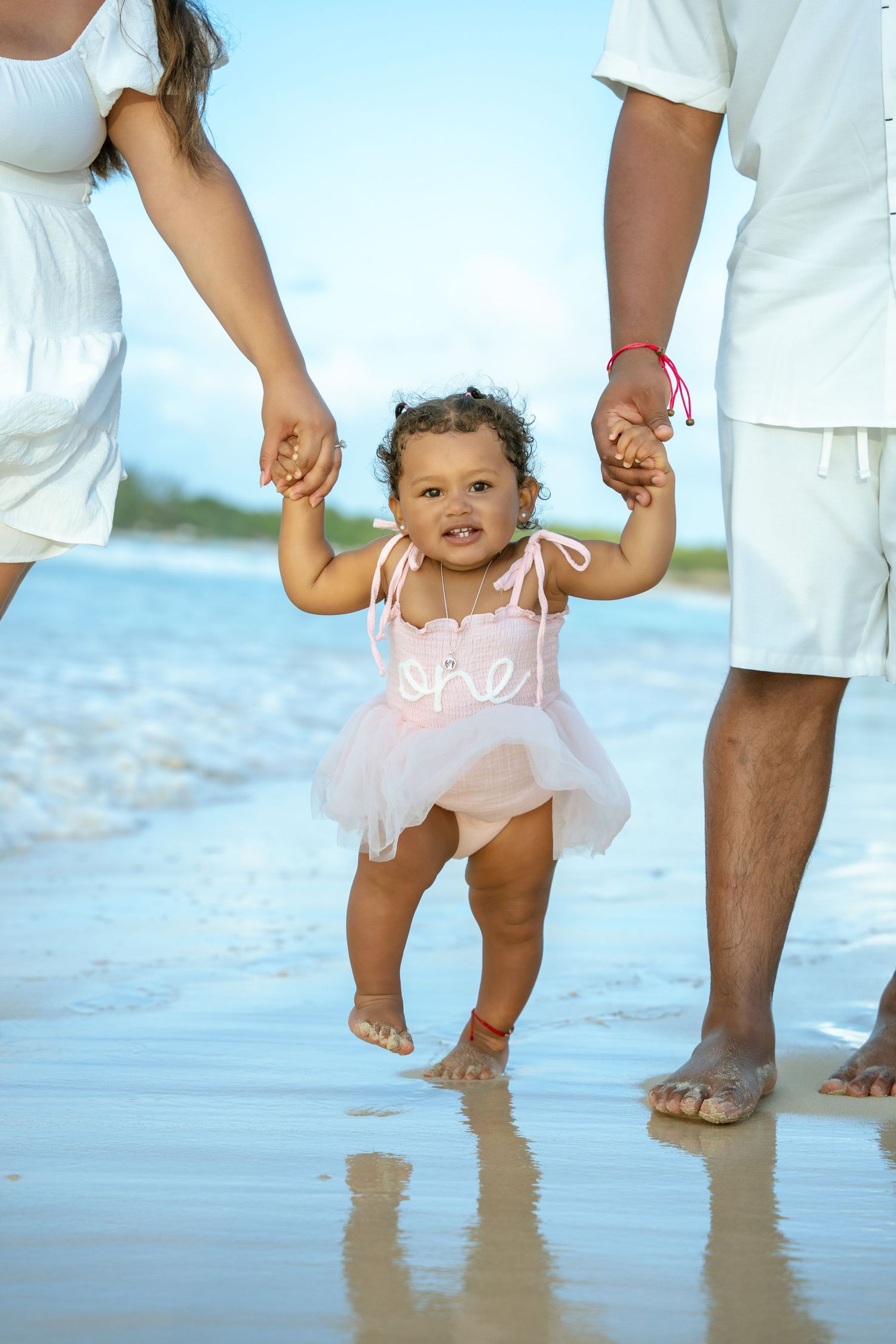 Toddler in a pink tutu outfit holds two adults' hands and takes steps on a wet sandy beach with gentle ocean waves behind.
