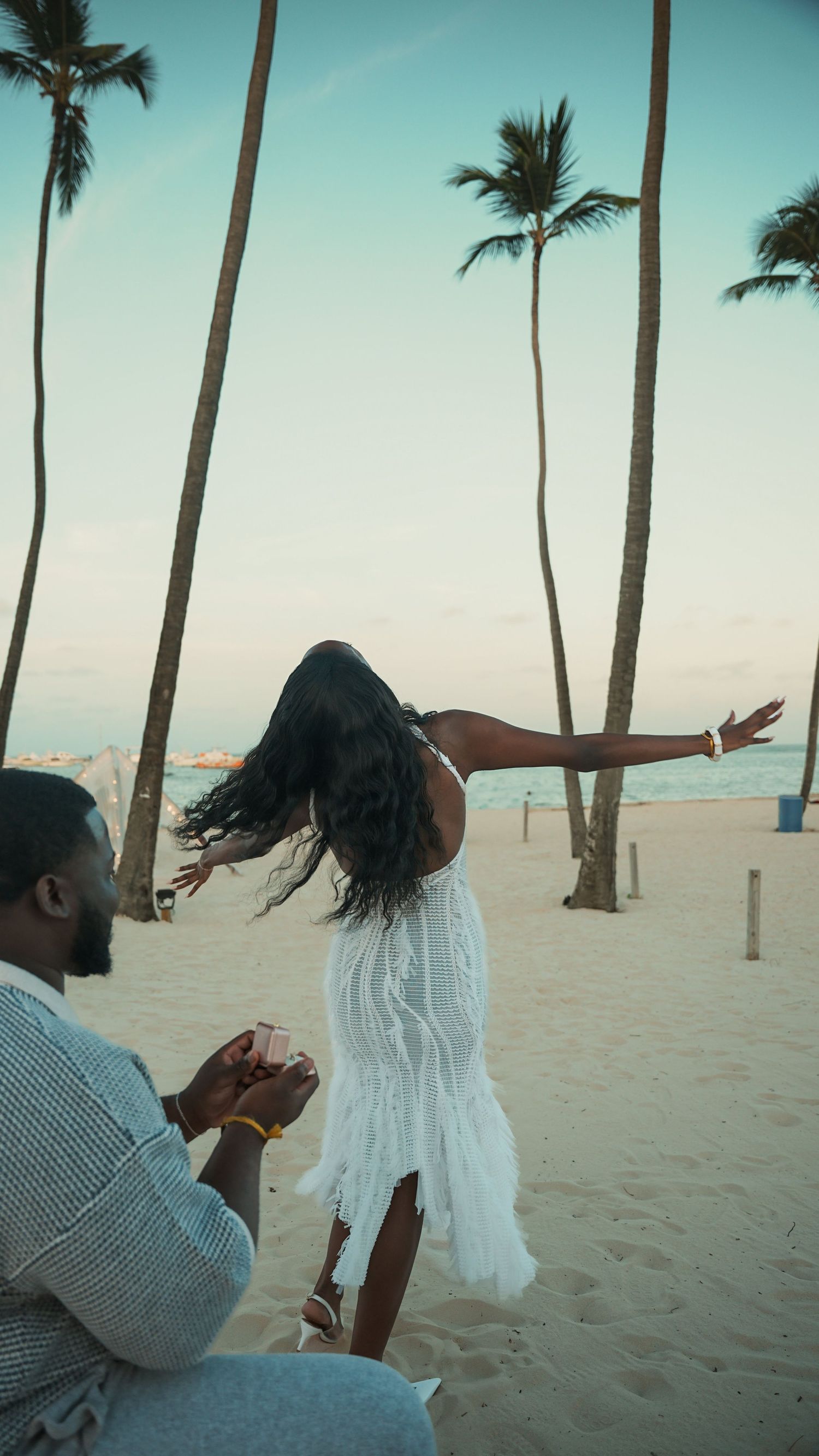 A man kneels on a tropical beach holding a ring box as a woman in a white dress spreads her arms near palm trees at sunset.