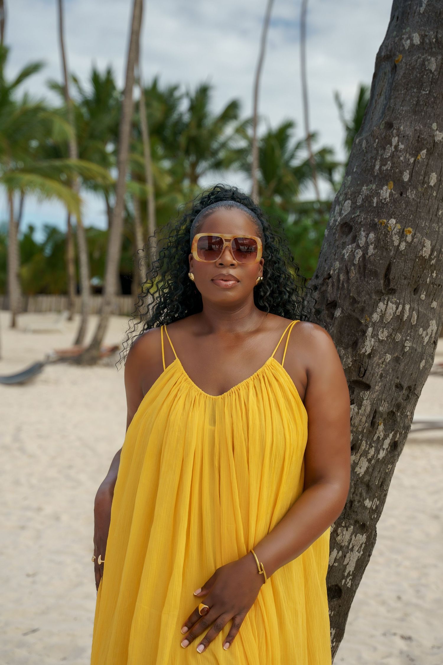 Woman in a flowing yellow sundress and tinted sunglasses leans against a palm tree on a sandy tropical beach.