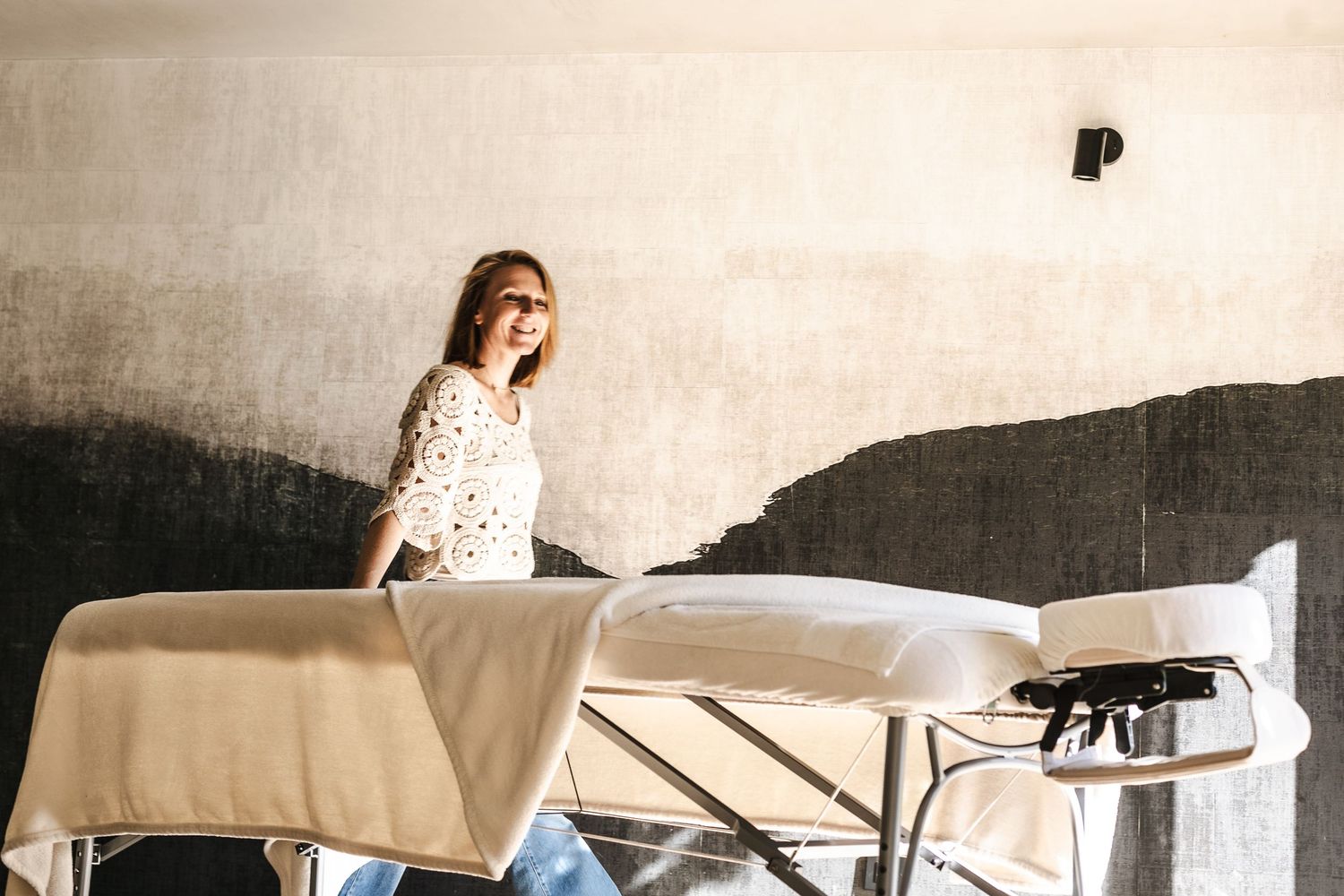 Praticienne souriante et lumineuse avec une main posée sur sa table de massage. Linge propre et doux. Ambiance de massage chaleureux.